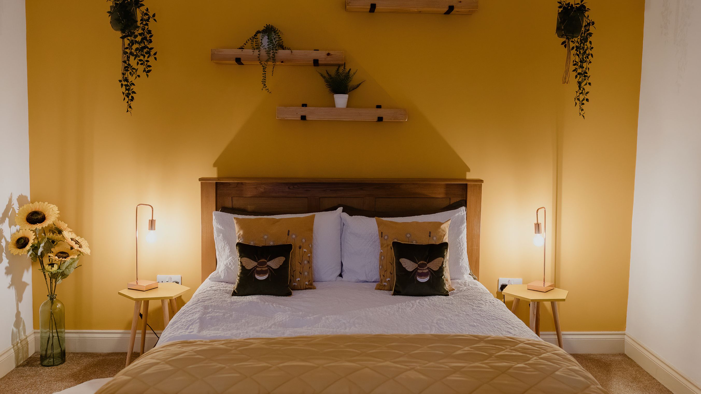 Cosy bedroom with mustard accent wall, wooden bed, bedside lamps and hanging plants