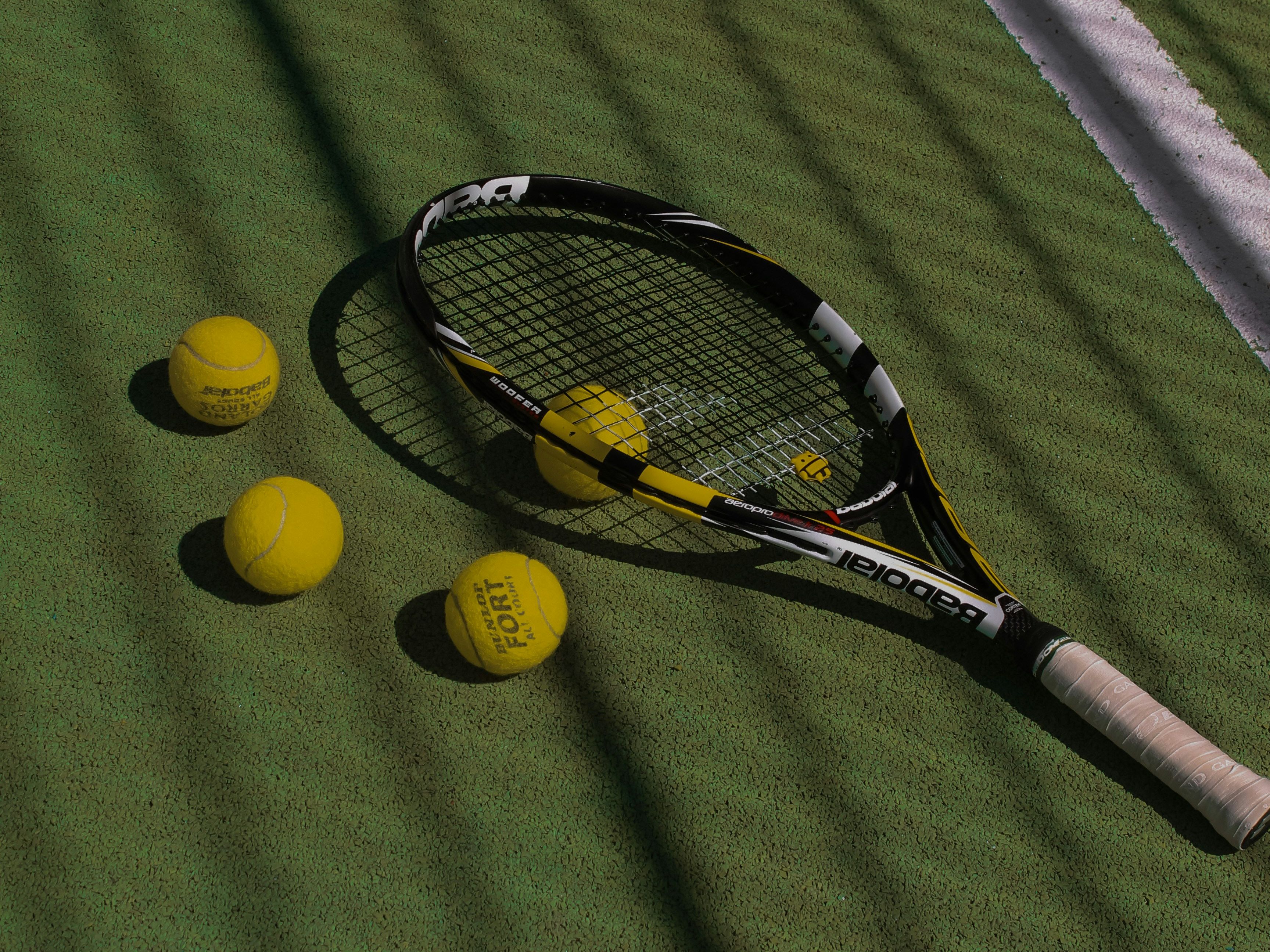Tennis racquet and three yellow tennis balls on a green court near a white line with fence shadows