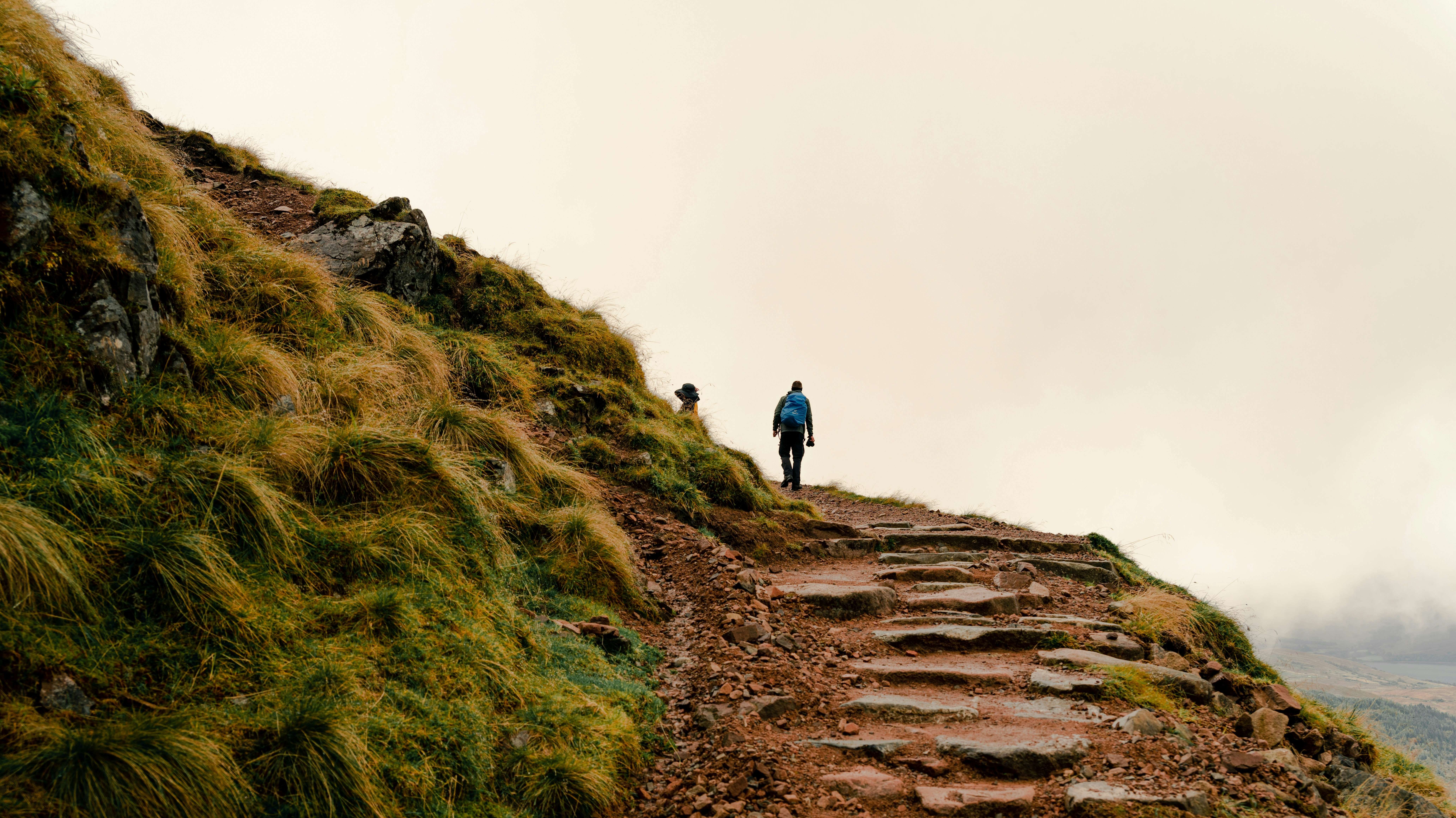 Hiker with a backpack walking up a rocky mountain path with stone steps beside grassy slopes under an overcast sky
