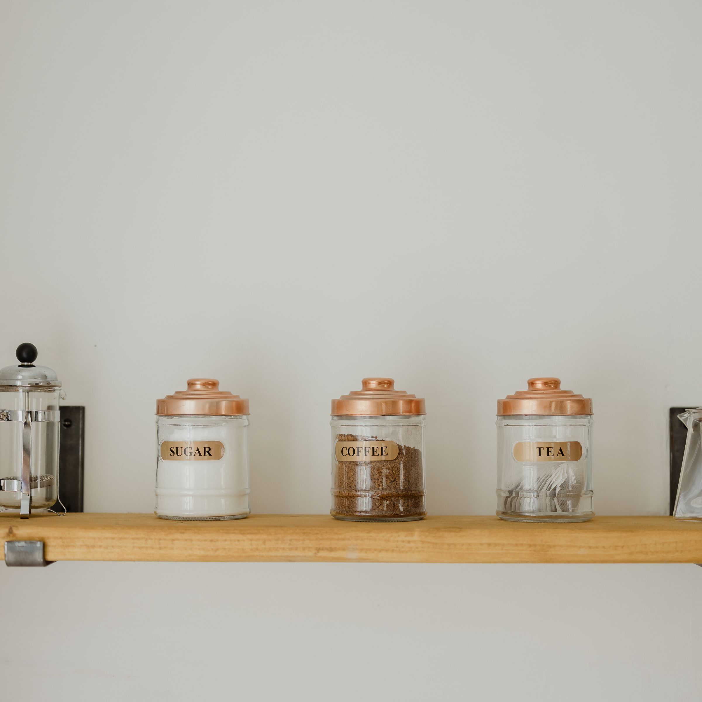 Glass jars labelled sugar, coffee and tea on a wooden shelf with a French press and a glass teapot