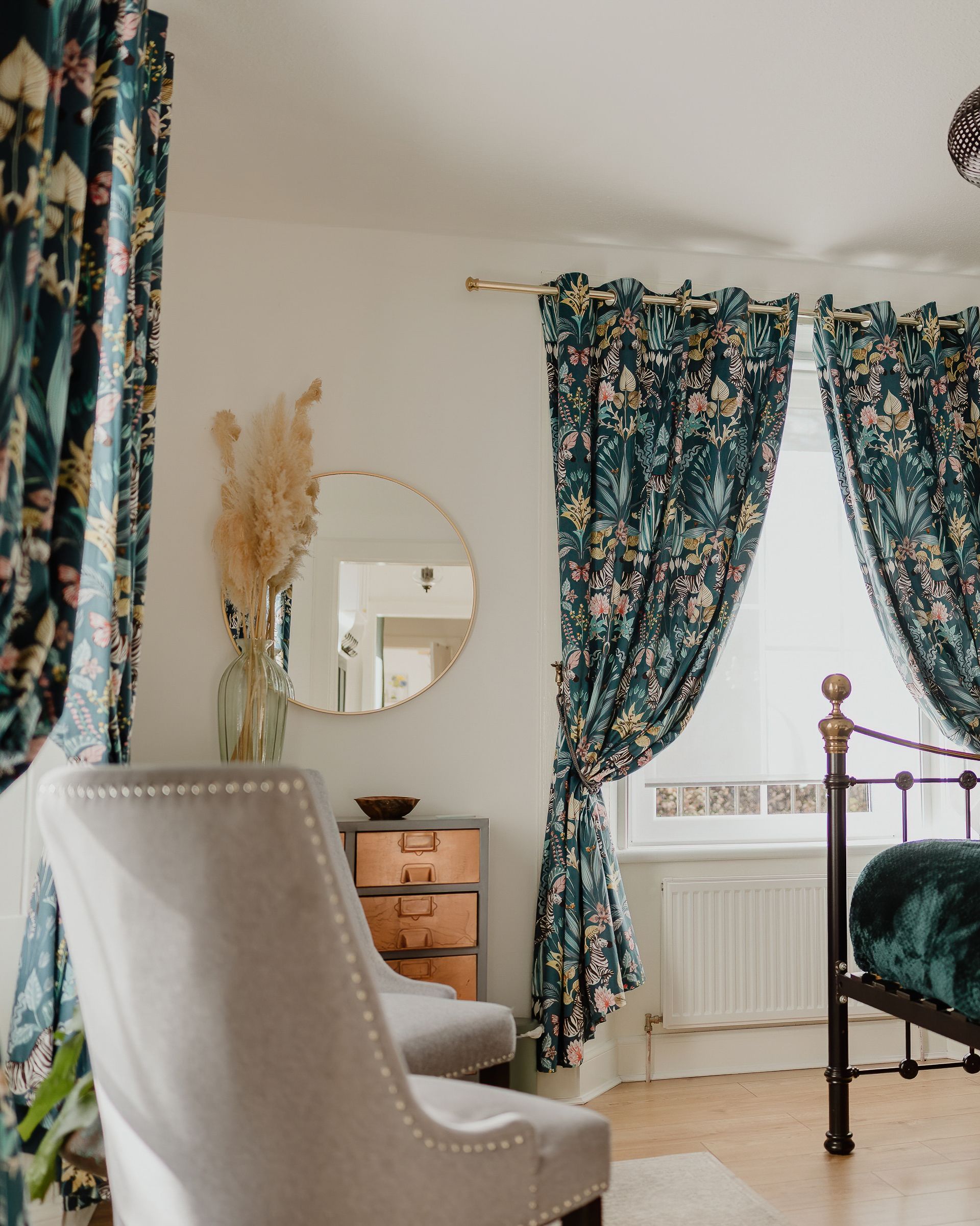 Bedroom with teal floral curtains, brass bed frame and a round wall mirror