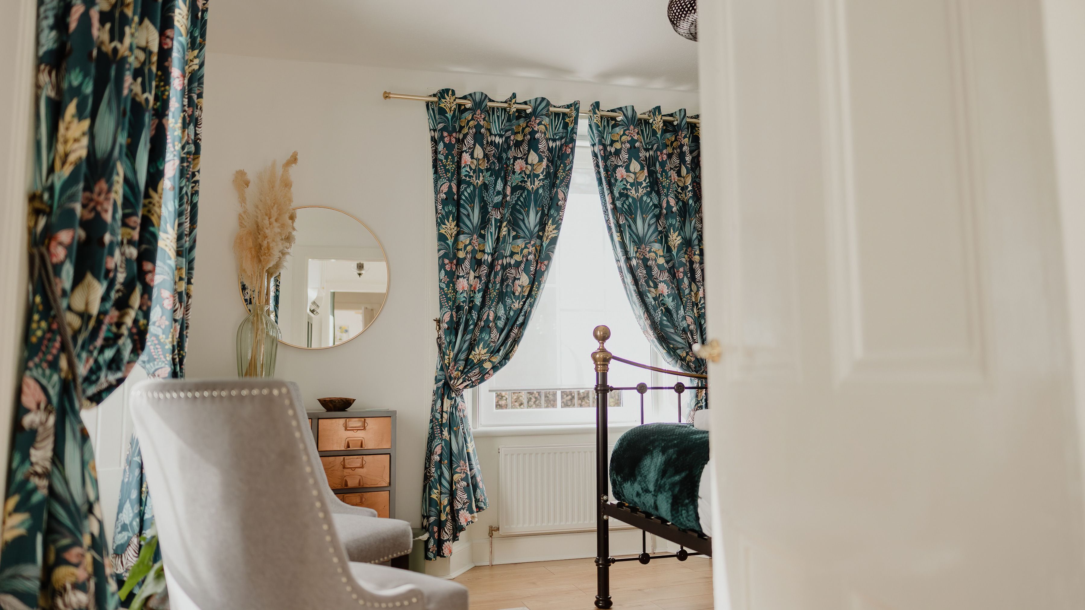 Bedroom with teal floral curtains, brass bed frame and a round wall mirror
