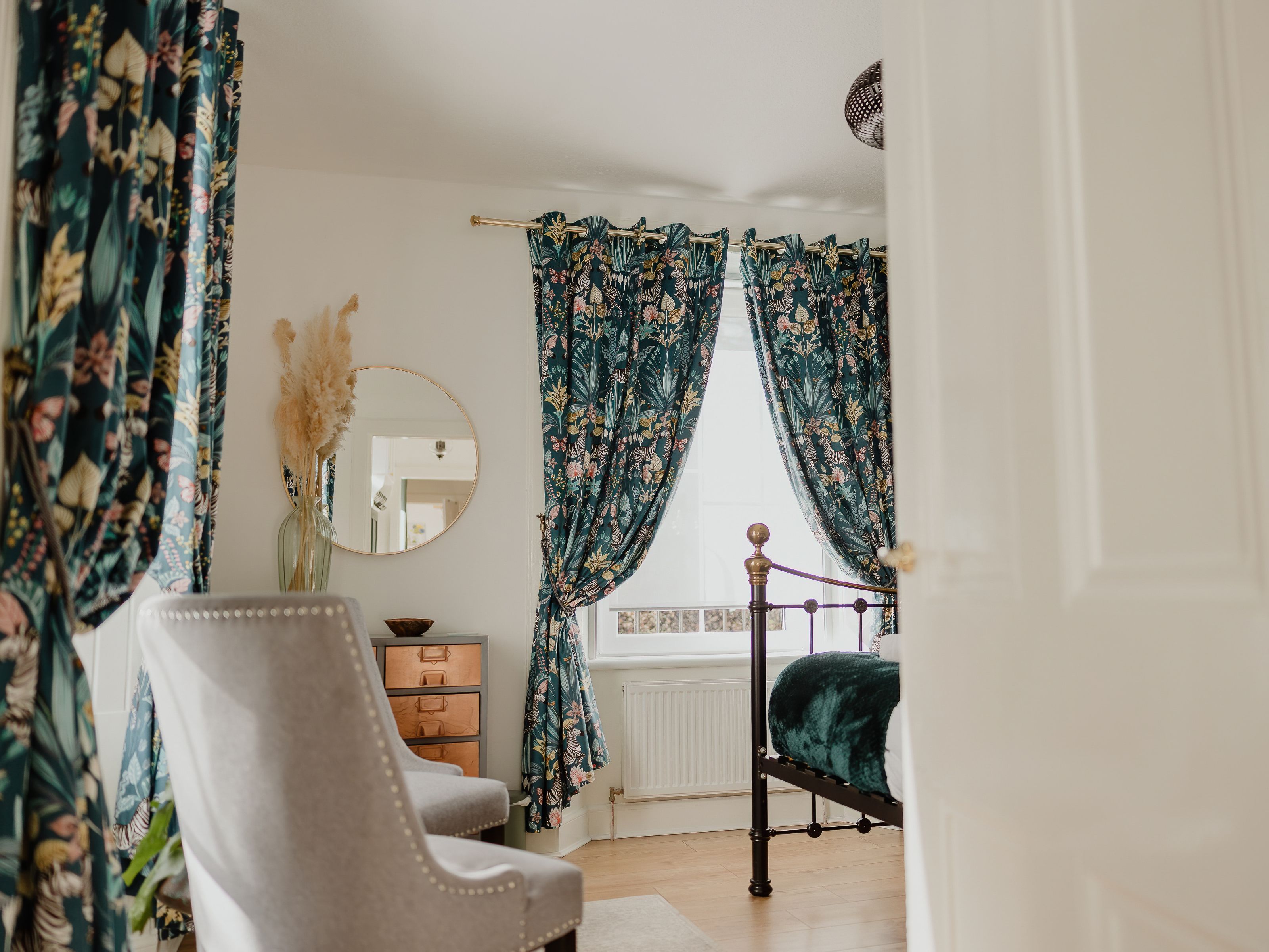 Bedroom with teal floral curtains, brass bed frame and a round wall mirror
