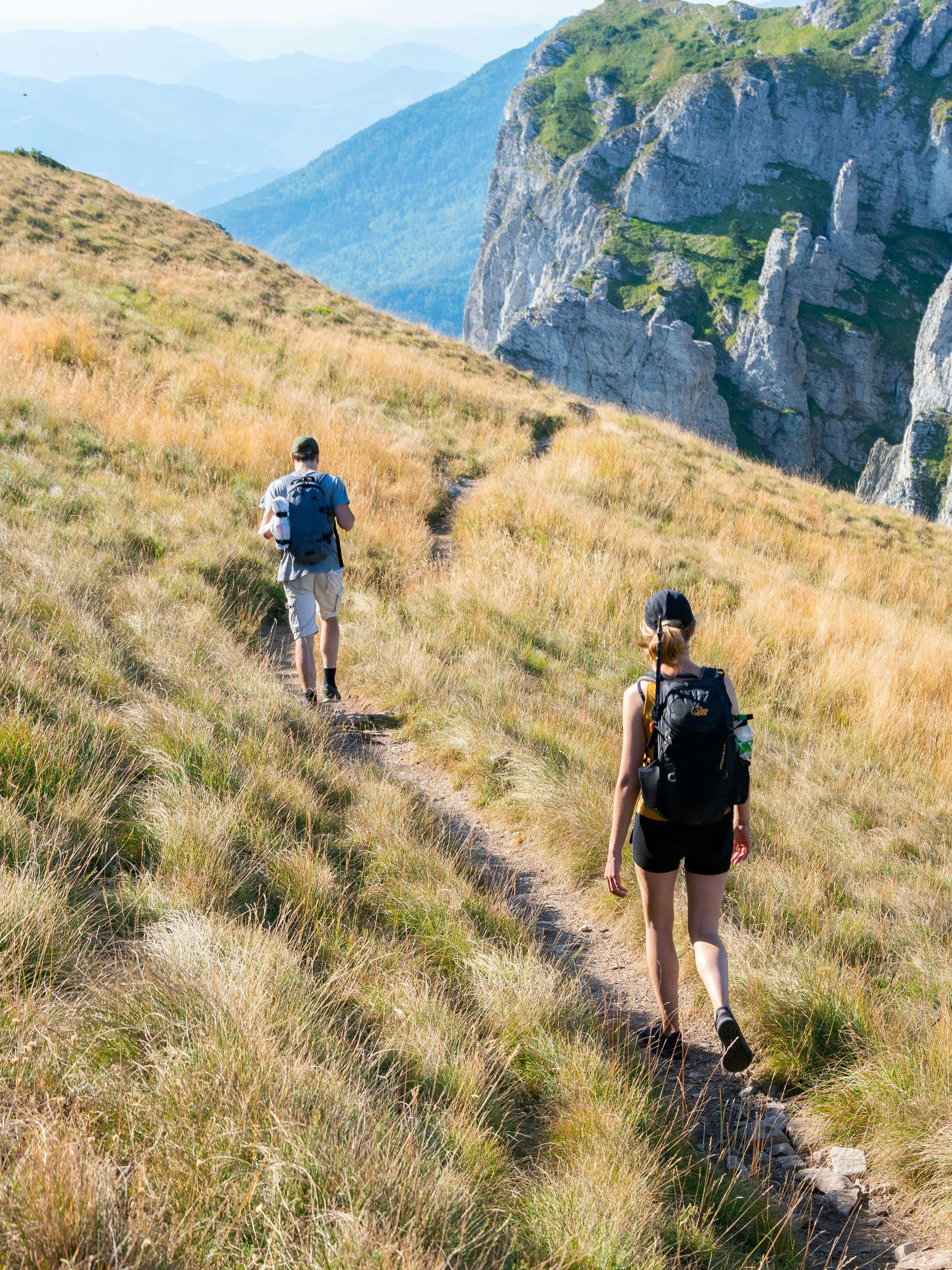 Two hikers walking along a narrow path on a grassy mountain ridge beside rocky cliffs