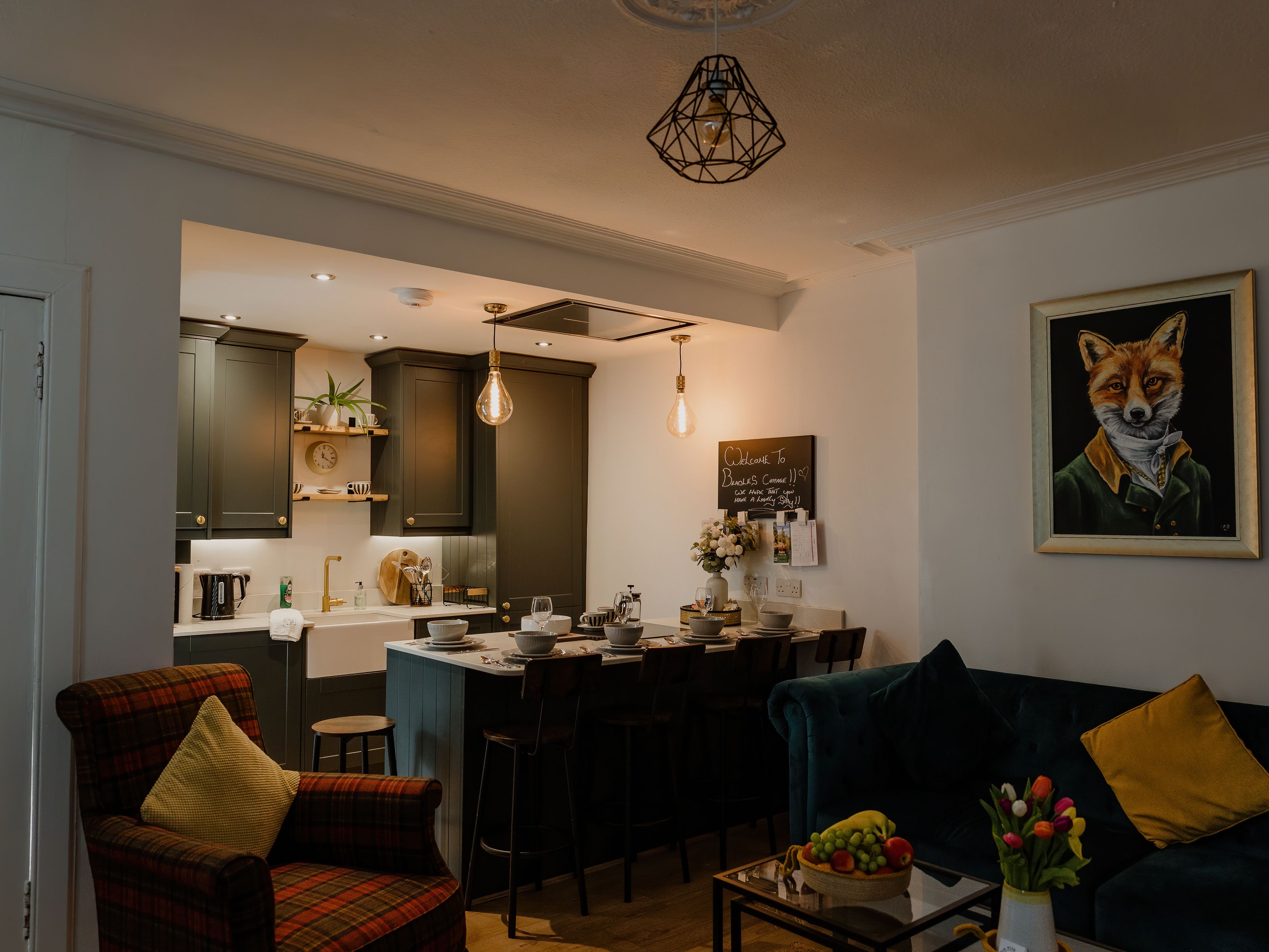 Cosy open-plan kitchen and dining area with dark cabinets, pendant lights and a set table beside a sofa