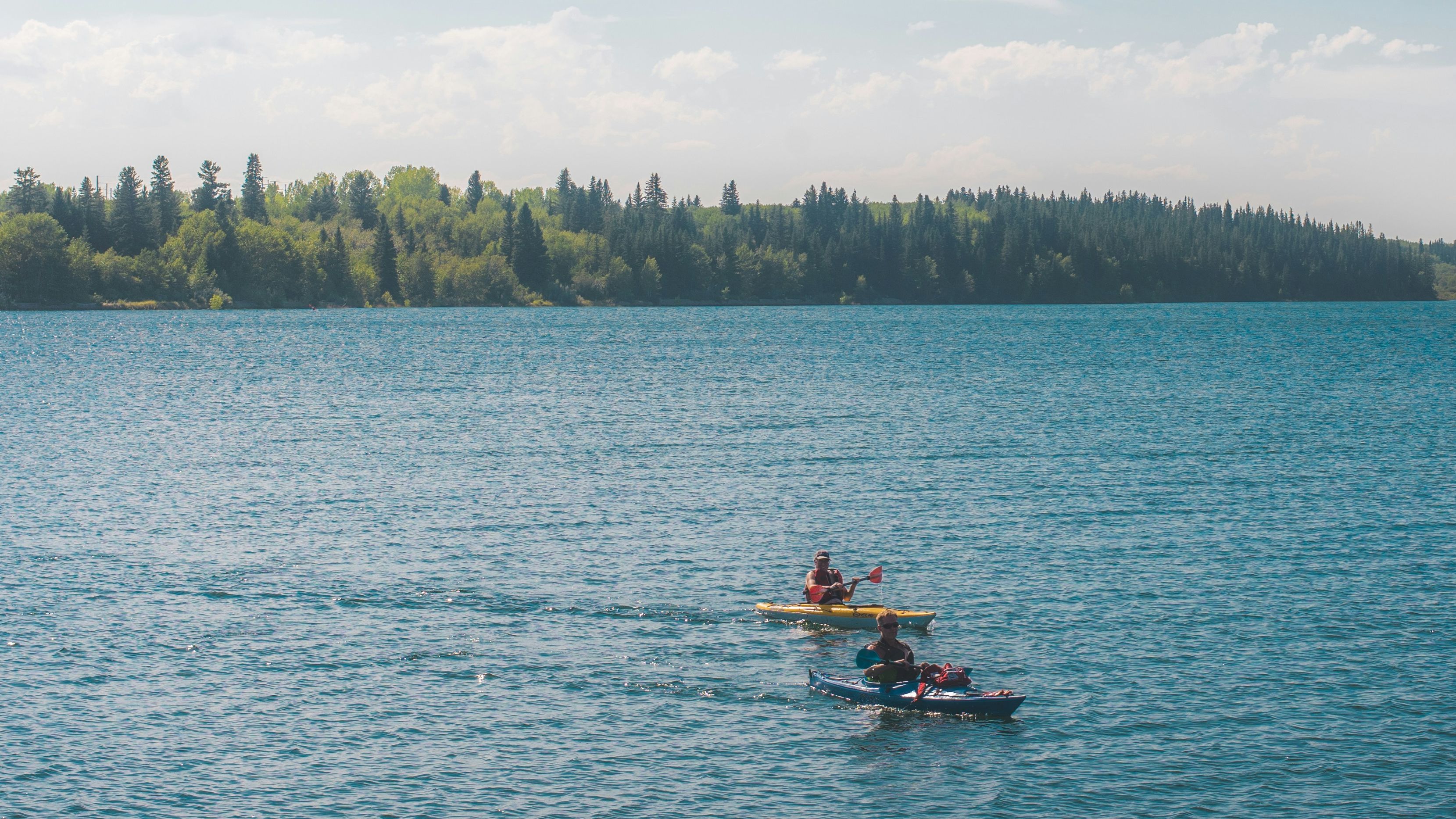 Two people kayaking on a calm blue lake with a forested shoreline under a wide sky.