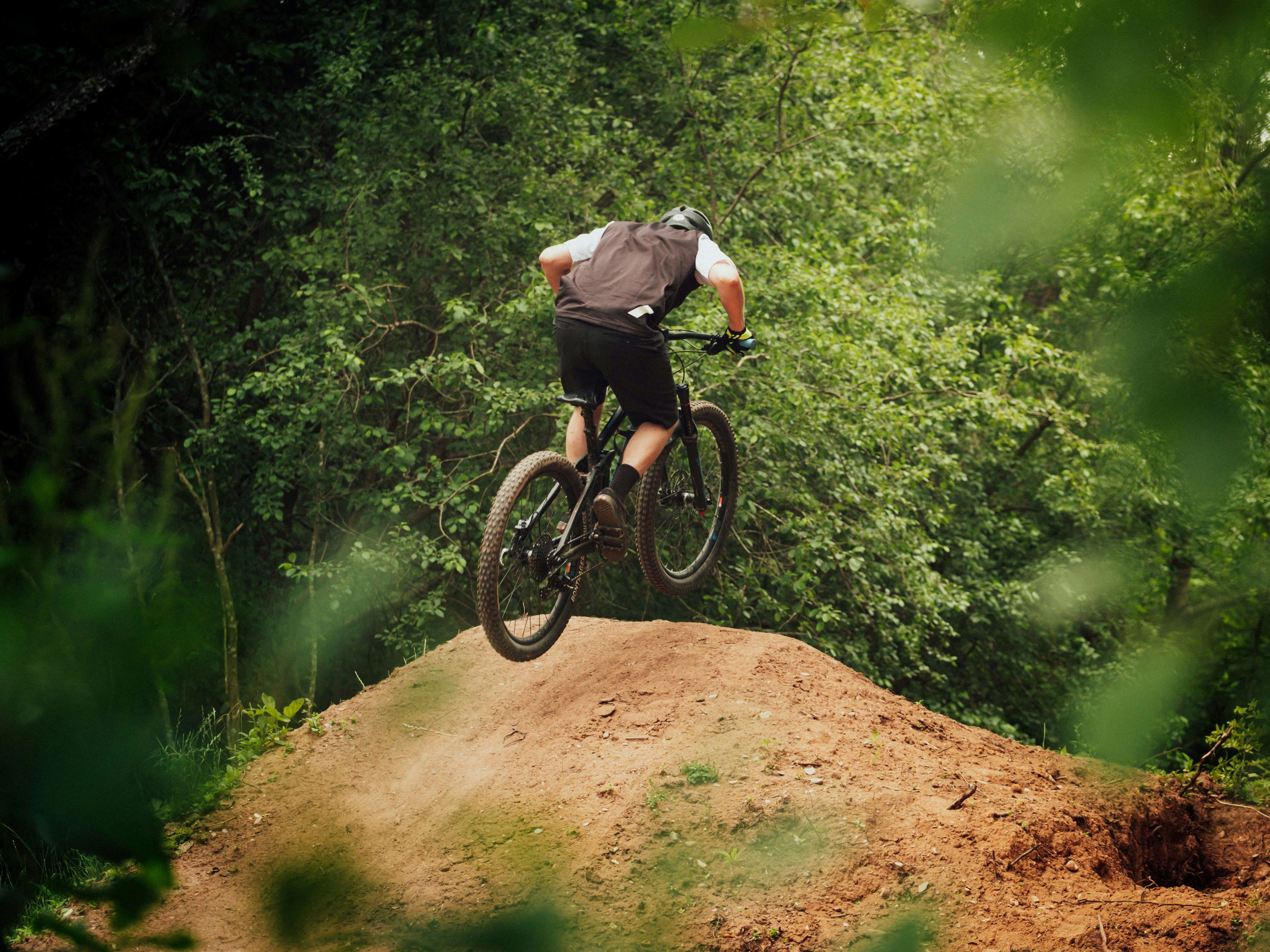 Mountain biker launching off a dirt jump in a green forest
