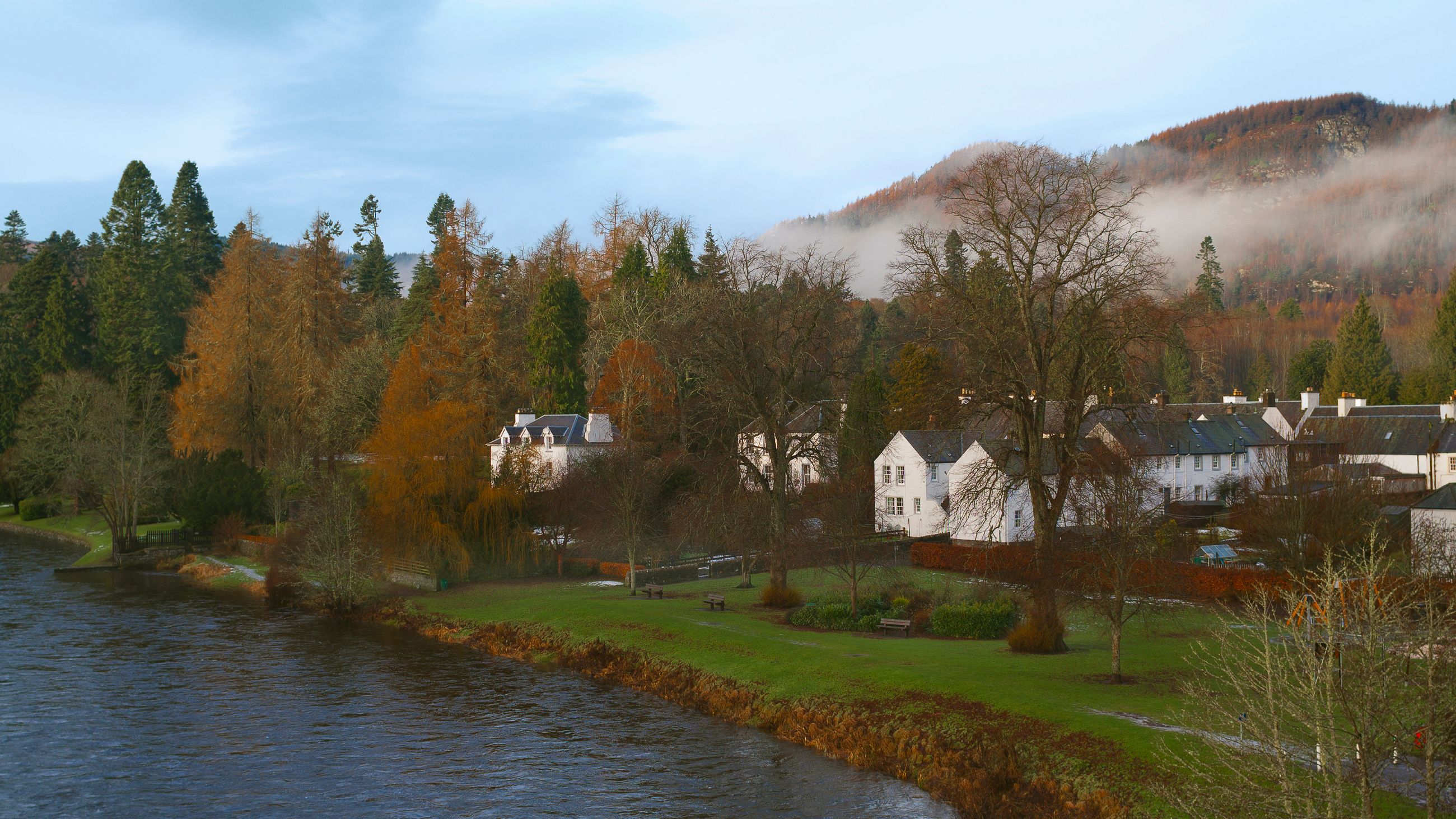 Riverside village with white houses, autumn trees and misty hills under a pale blue sky