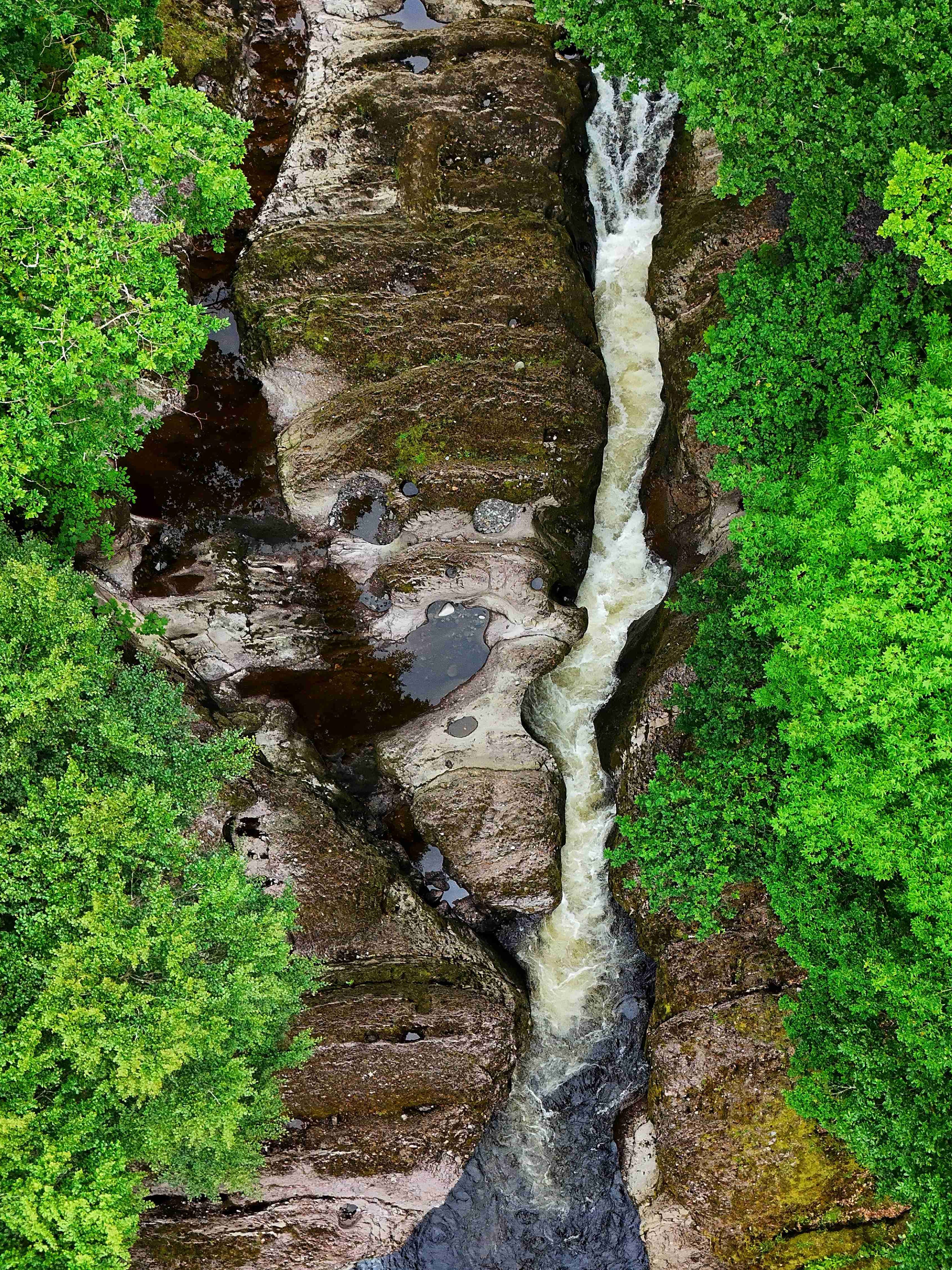 Aerial view of a narrow waterfall cutting through rocks in a dense green woodland