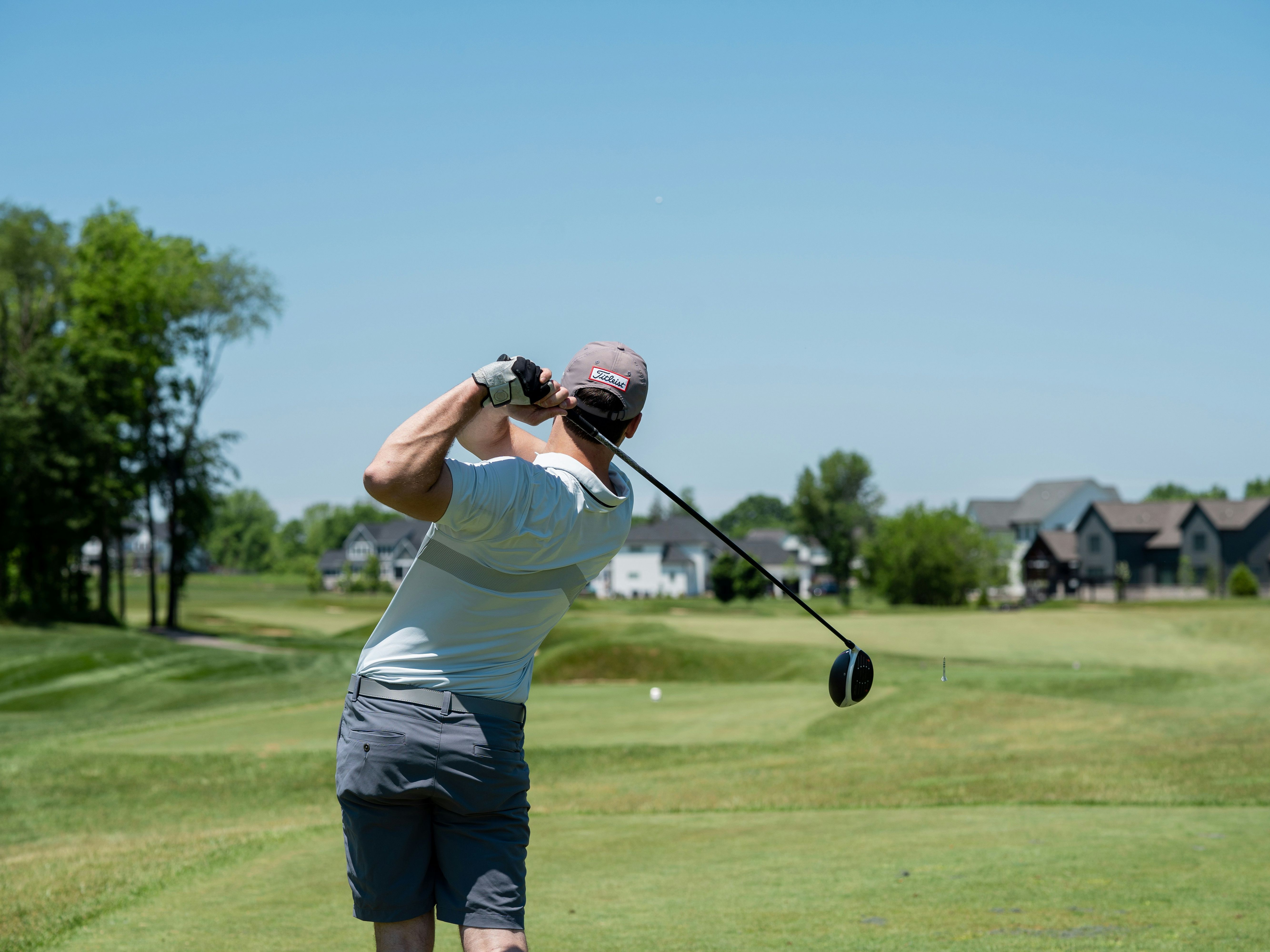 Golfer finishing a drive on a sunny course with houses in the distance