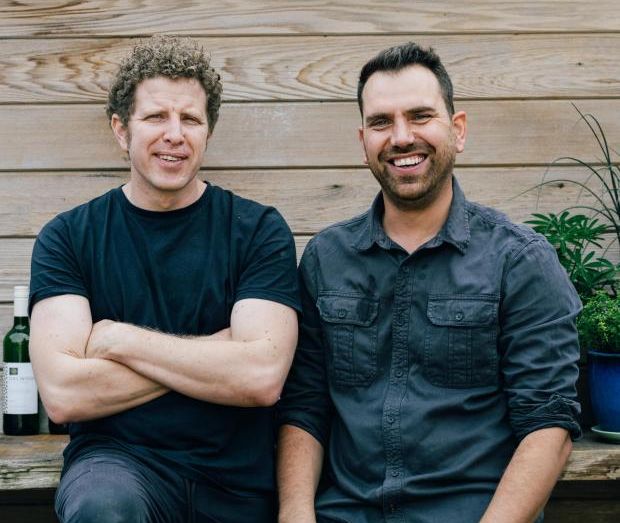 Two men sitting together in front of a wooden wall with potted plants on a shelf above them.
