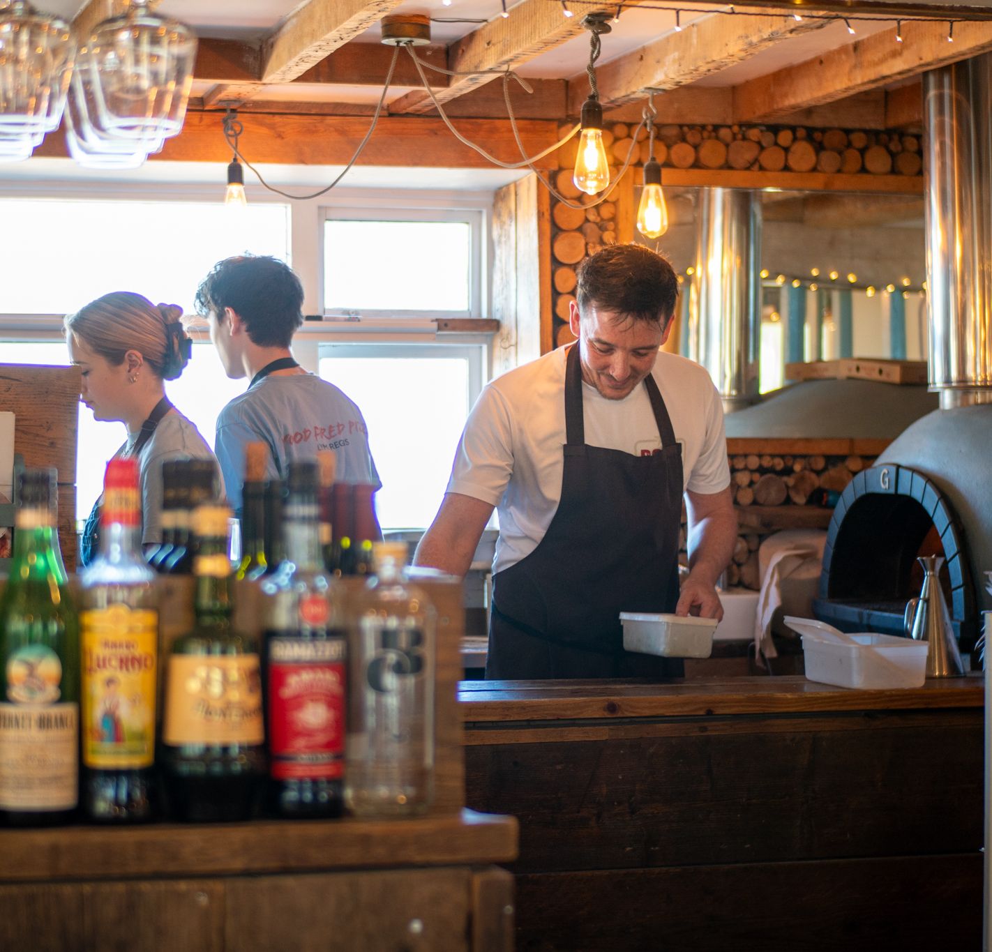 Staff working behind the bar in a rustic pizzeria with bottles in the foreground and a wood-fired oven in the background