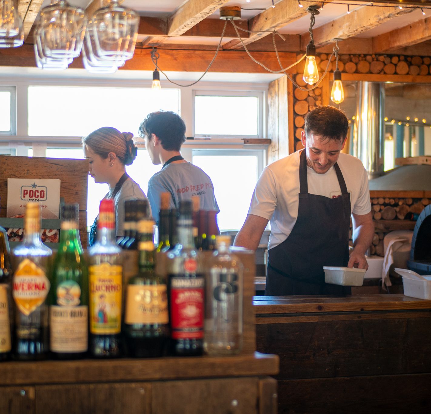 Staff working behind the bar in a rustic pizzeria with bottles in the foreground and a wood-fired oven in the background