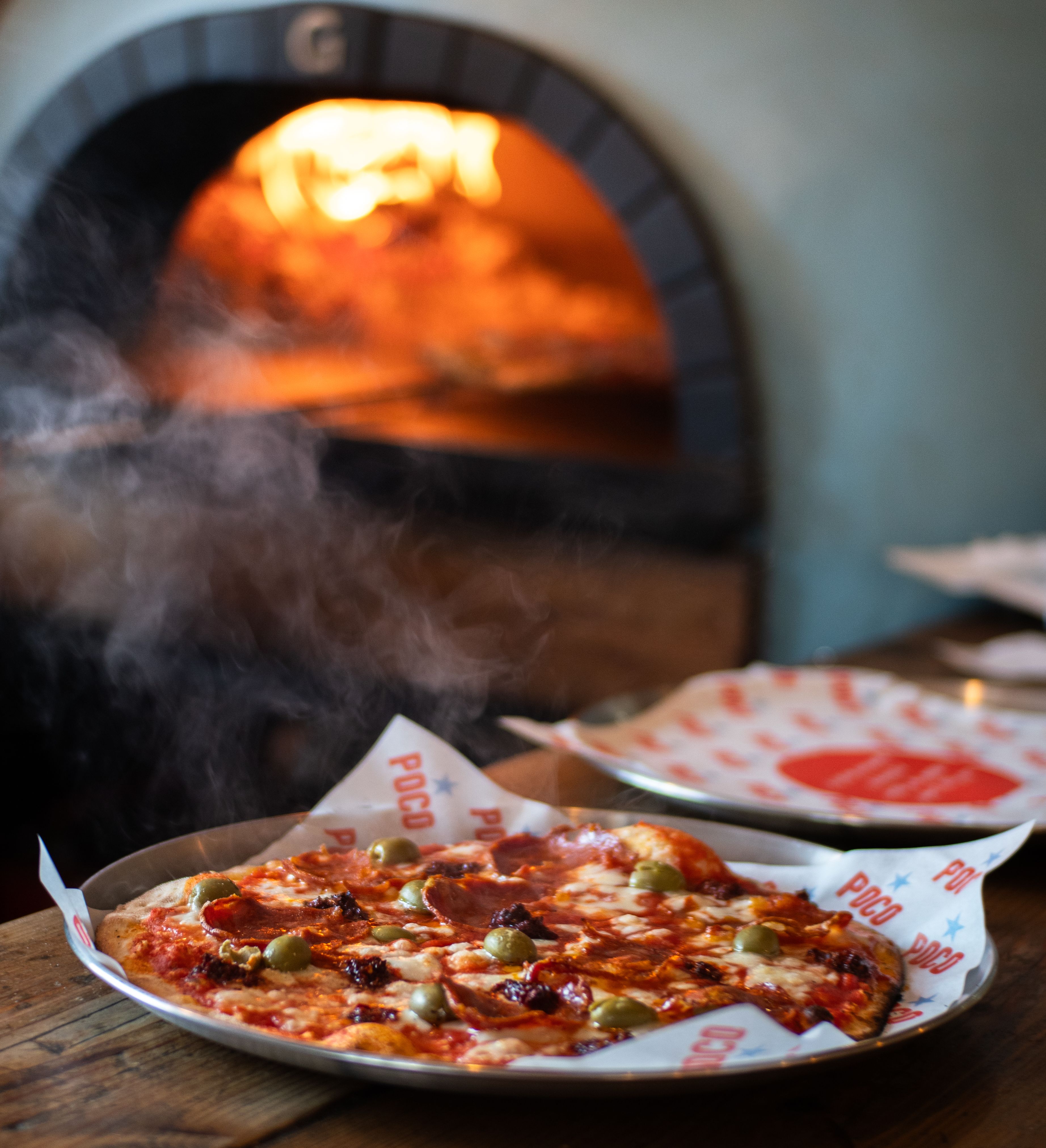 Freshly baked pizza on a metal tray with steam rising, in front of a glowing wood-fired oven