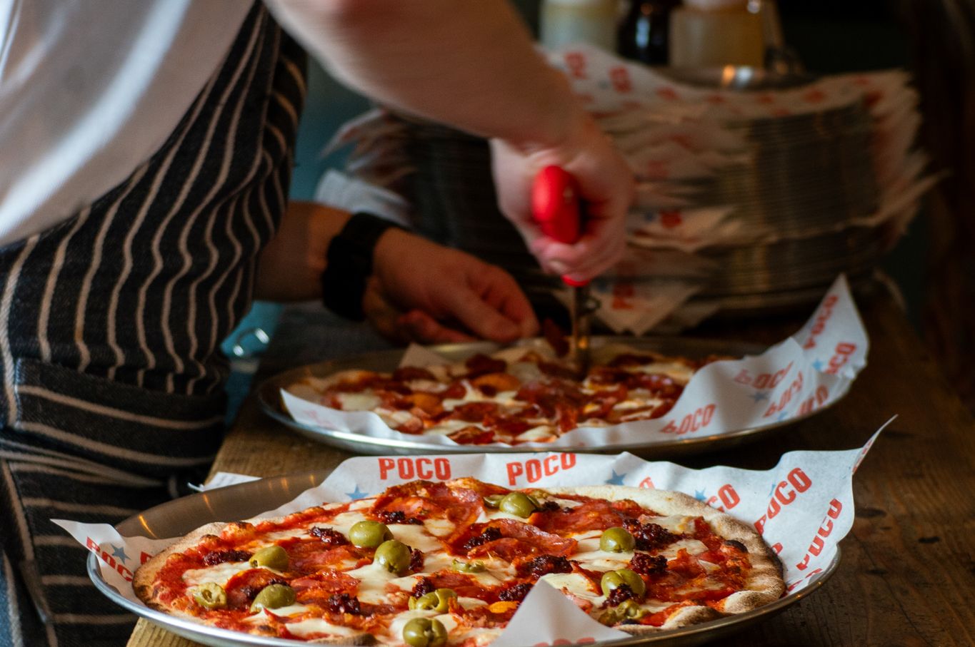 Two pizzas on the pass with bowls of slaw, as a staff member cuts one with a pizza wheel