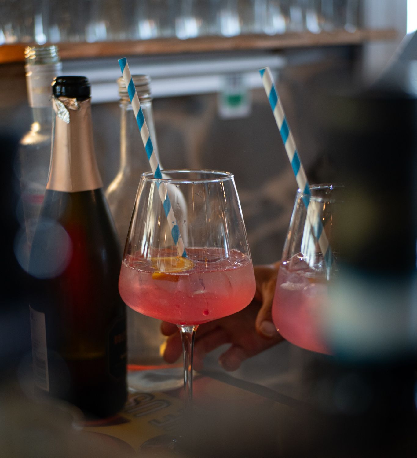 Two pink spritz cocktails in stemmed glasses with blue-and-white striped straws on a bar counter