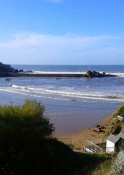 View of a beach with gentle waves, small boats, rocky outcrops, green hills, and small white beach huts under a clear blue sky.