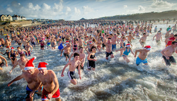 Large group of people participating in a festive beach swim, many wearing Santa hats and swimwear.