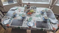 Dining table set for four with plates, cups, cutlery, and a centerpiece of fruit and condiments.