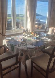 A dining table set for four with sunlight streaming through large windows, overlooking a coastal view.