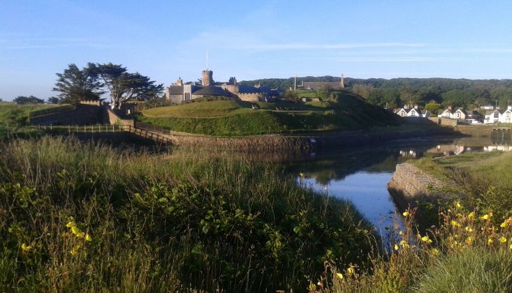 A scenic view of a historic castle-like building on a grassy hill with a moat and wildflowers in the foreground.