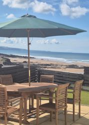 Outdoor wooden table and chairs with a large umbrella on a patio overlooking a sandy beach and ocean.