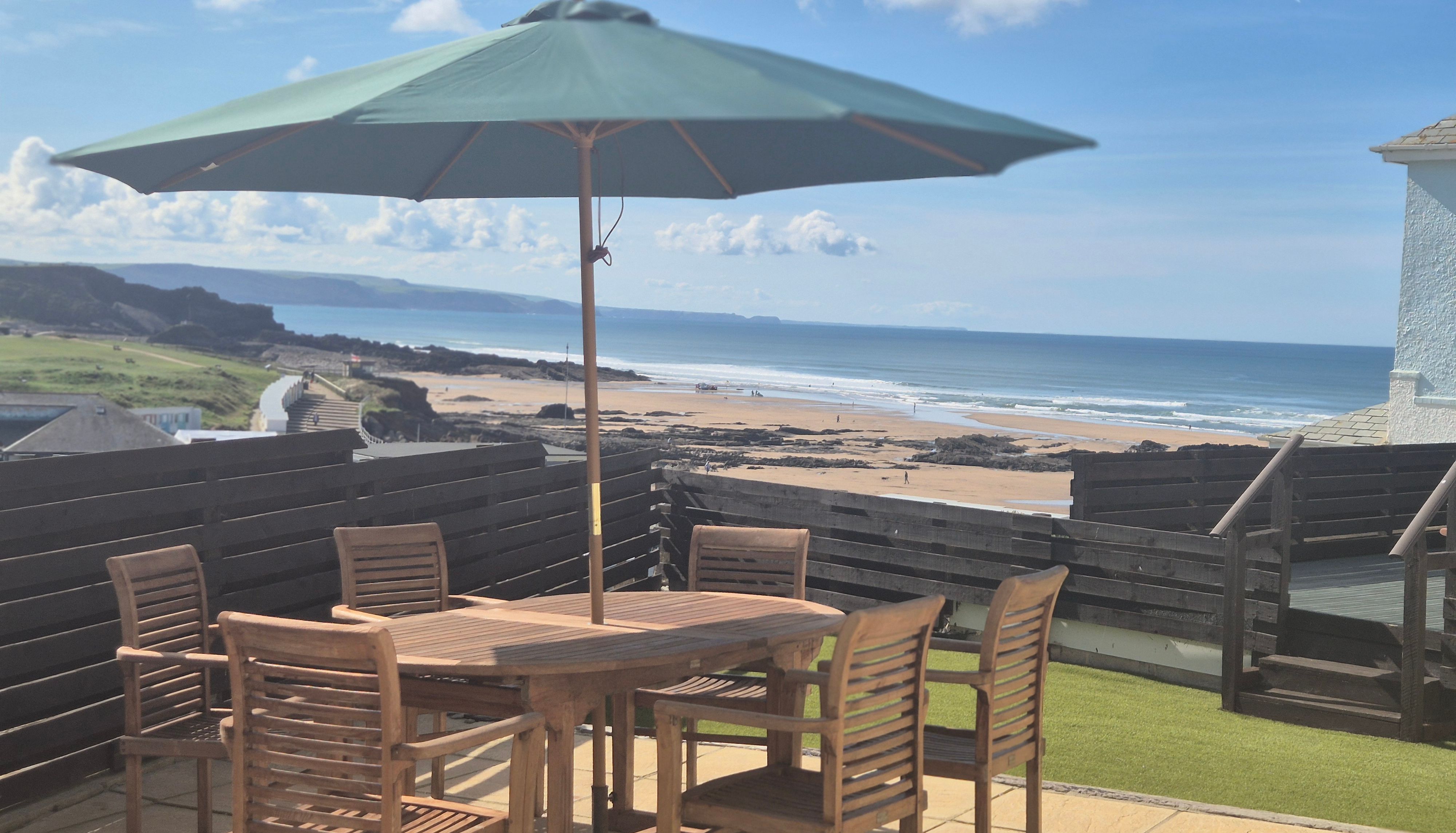 Outdoor wooden table and chairs with a large umbrella on a patio overlooking a sandy beach and ocean.