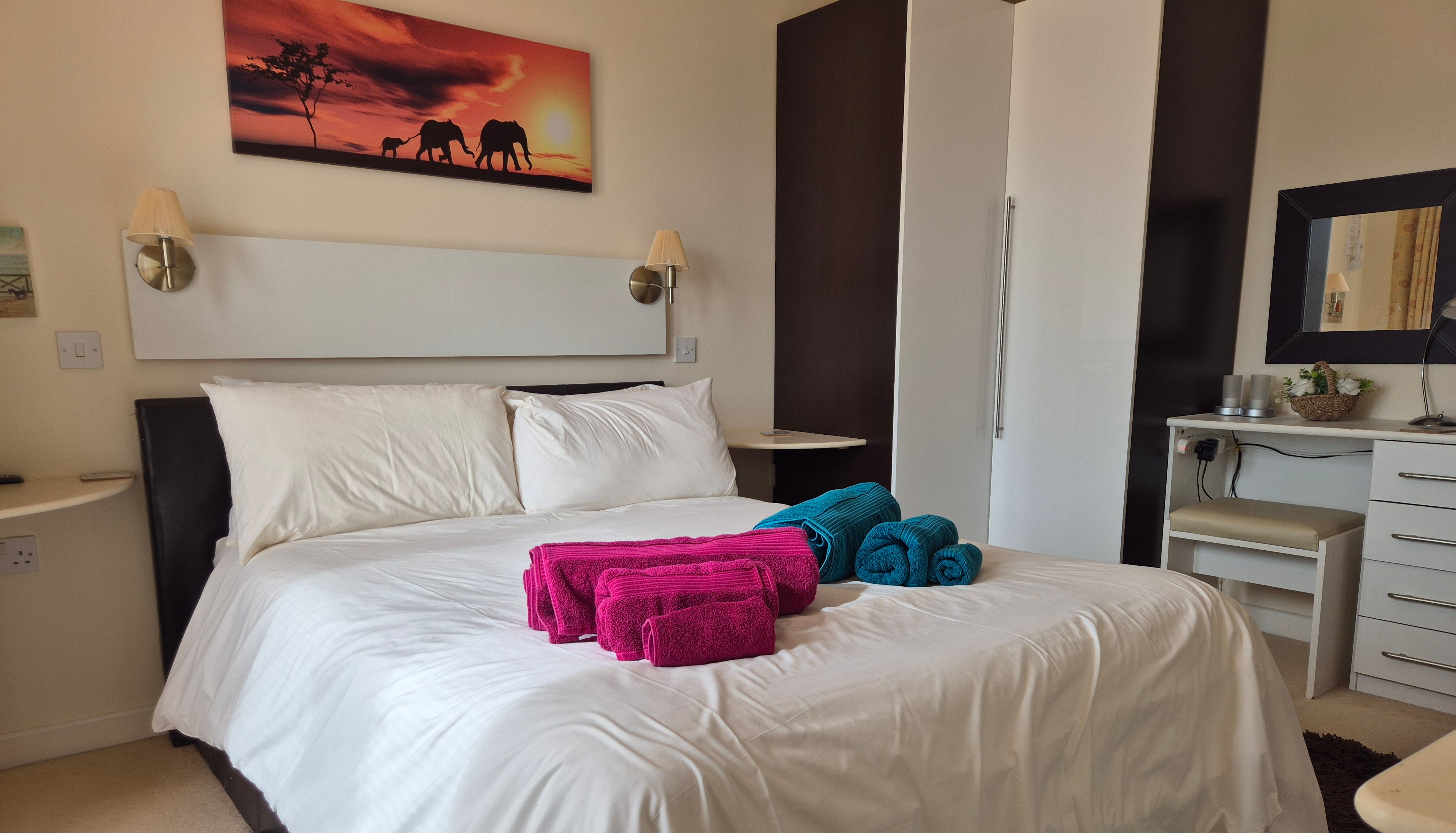 Modern bedroom with white bedding, colorful towels on the bed, and elephant wall art.