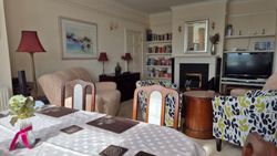 A cozy living room and dining area with patterned armchairs, a dining table with a white cloth, a TV, bookshelves, and modern decor.