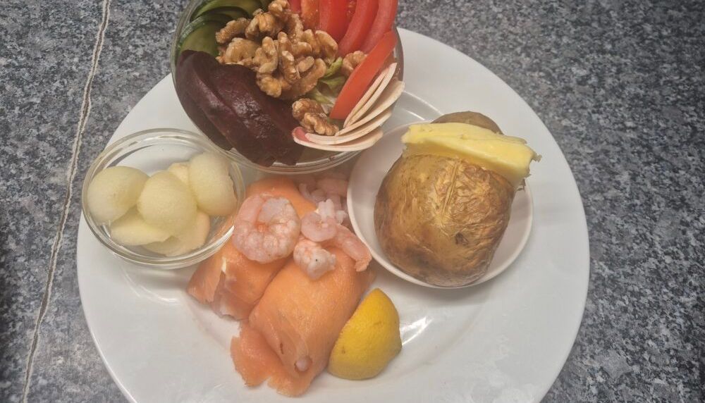 A plate of assorted cold foods including smoked salmon, prawns, baked potato with butter, salad with cucumber, tomato, beetroot, walnuts, and a small bowl of melon balls.