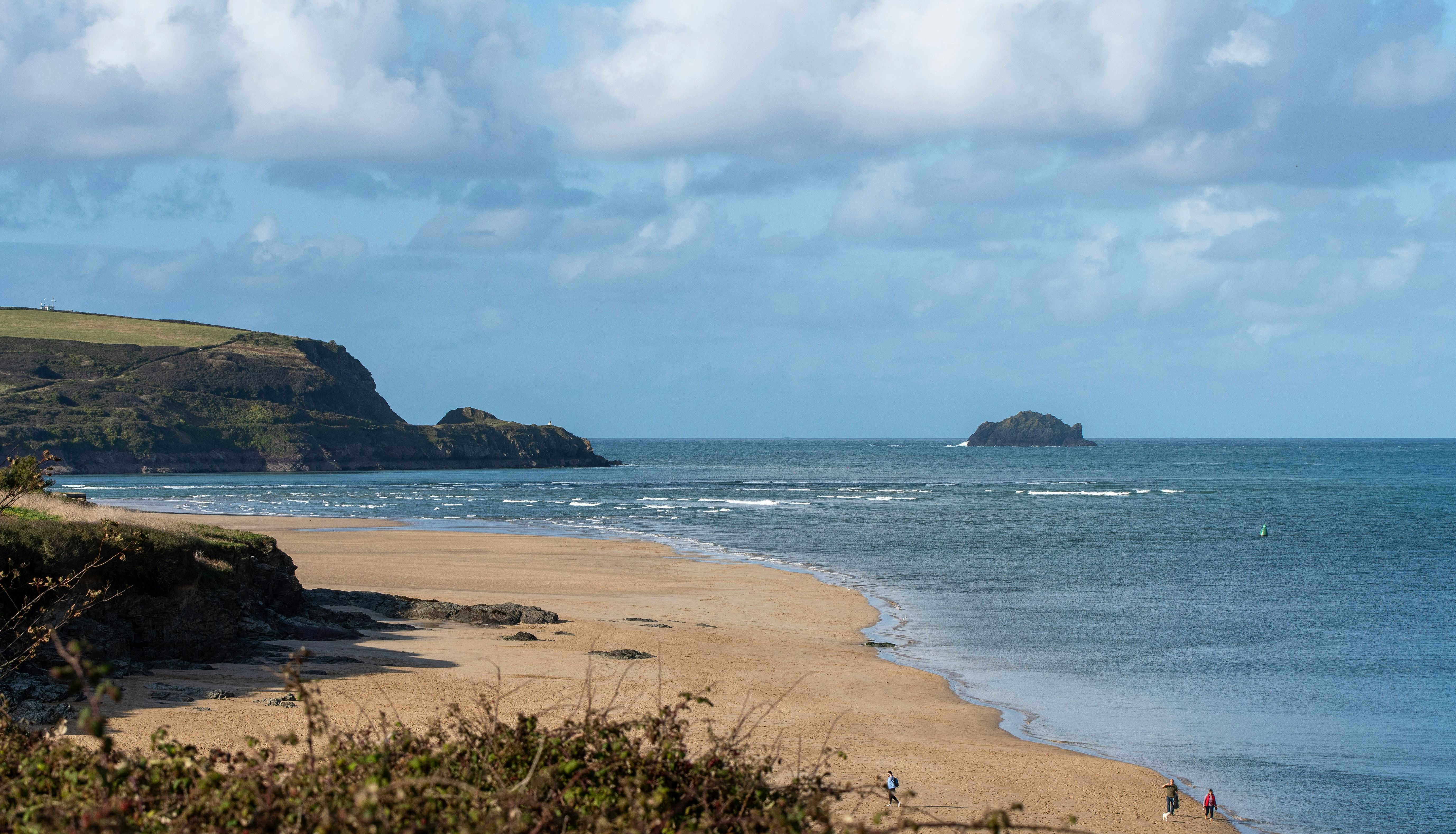 Scenic coastal landscape with sandy beach, rocky cliffs, and people walking along the shore.