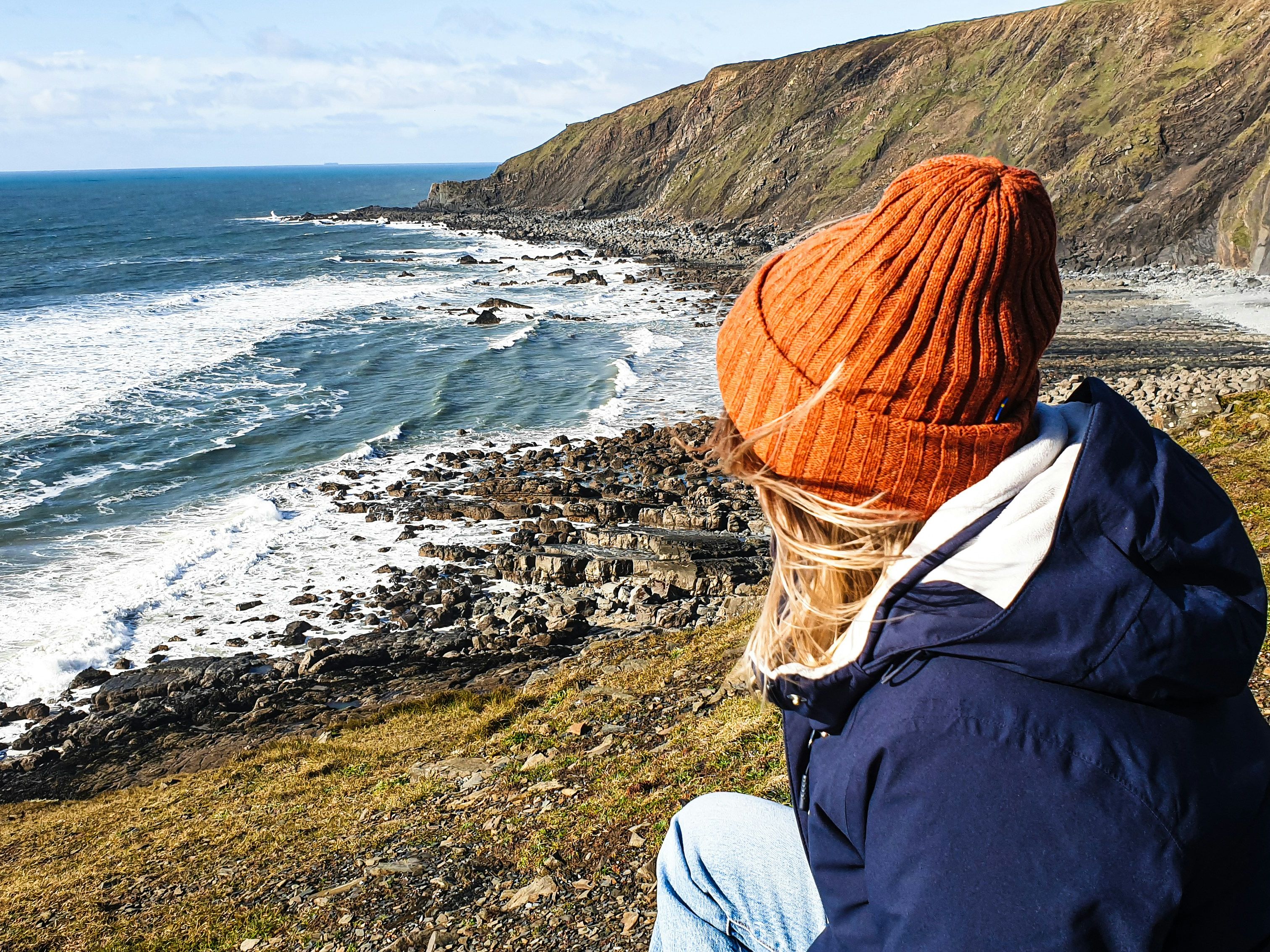 Person in orange beanie and blue jacket sitting on a grassy cliff overlooking a rocky coastline and ocean waves.
