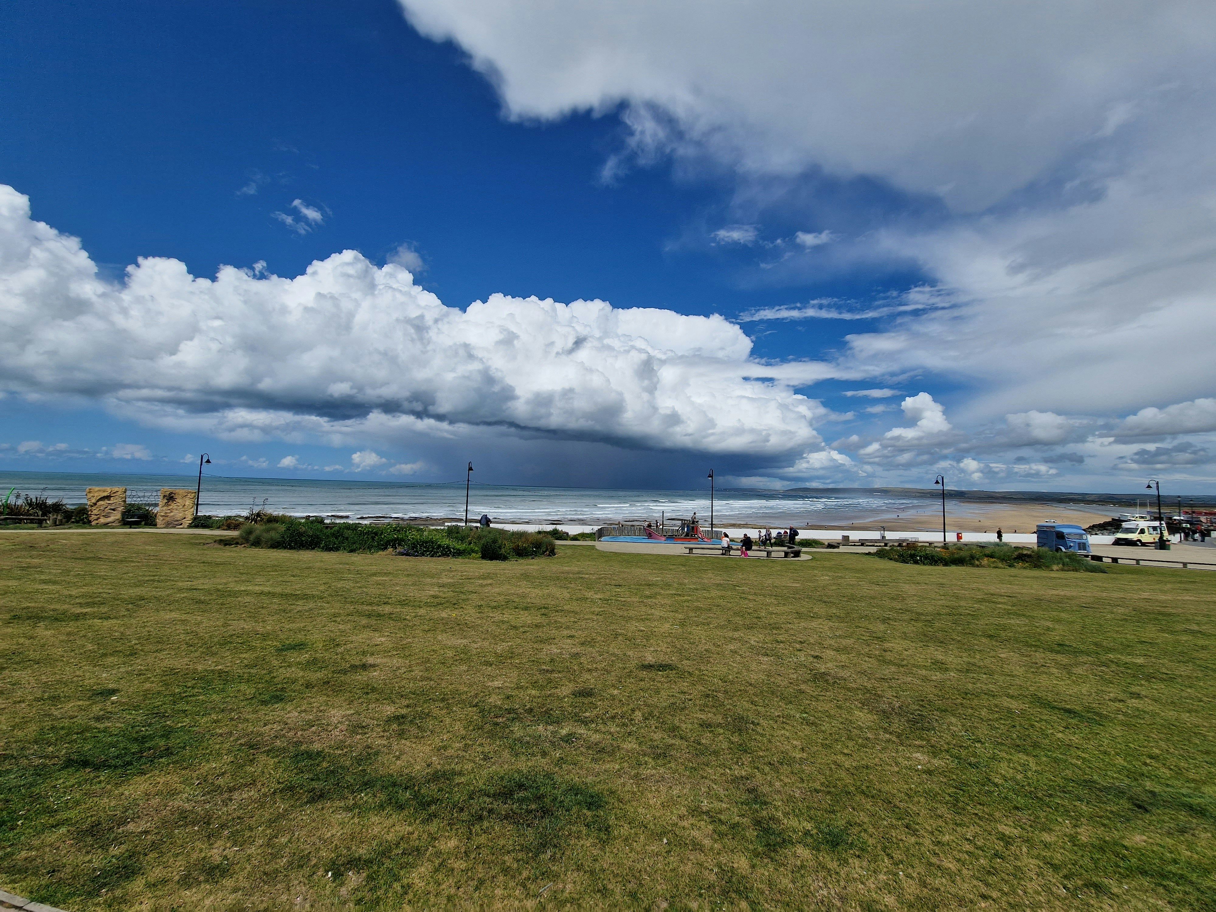 A coastal scene with a grassy park in the foreground, beach and ocean in the background, and large dramatic clouds in the sky.