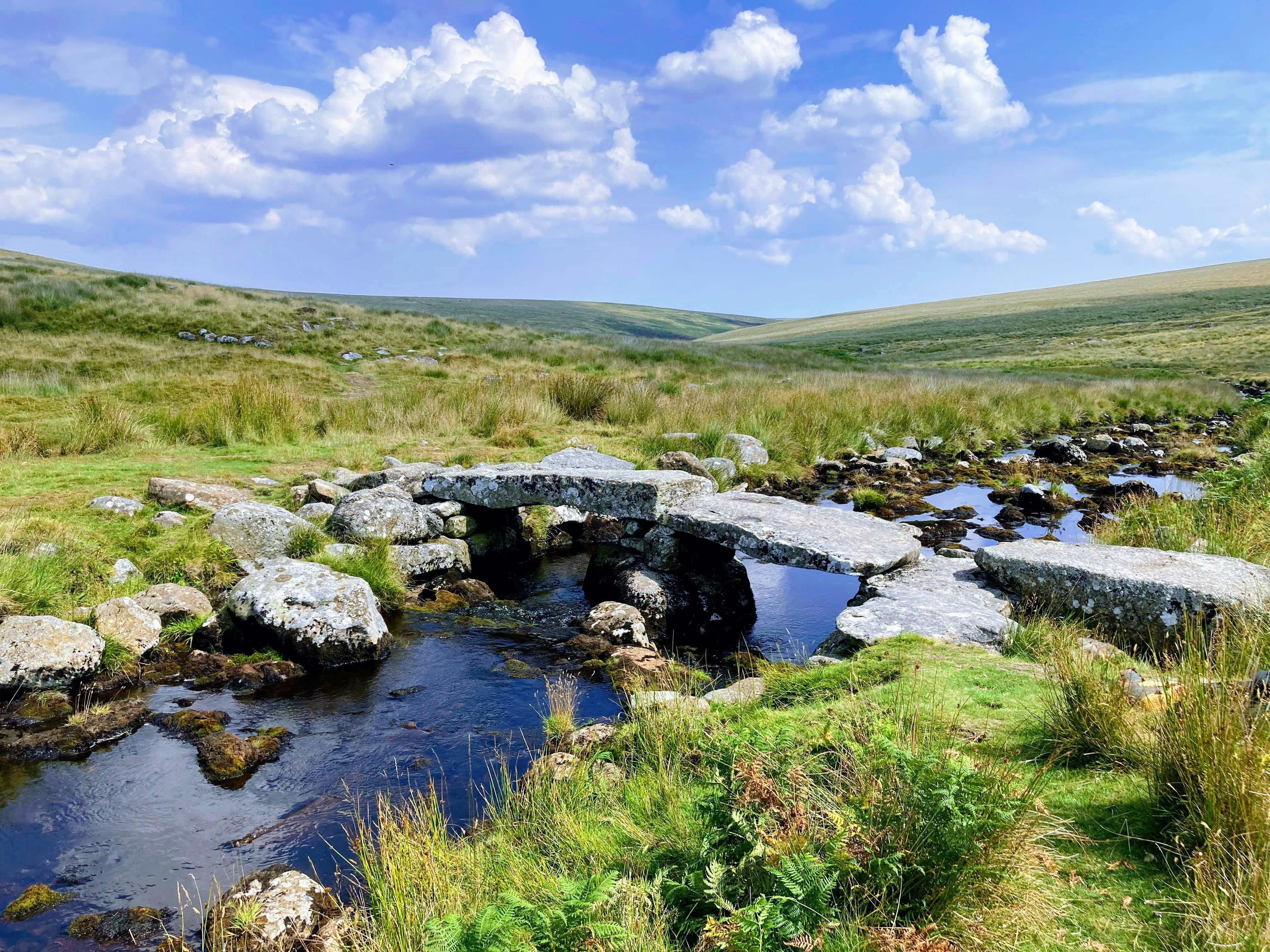 Ancient stone clapper bridge crossing a small stream in a grassy, open moorland landscape under a bright blue sky with scattered clouds.