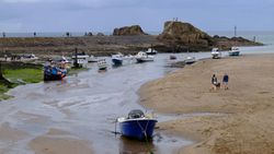 Boats resting on sand and shallow water at low tide near a rocky shore, with people walking and a cloudy sky in the background