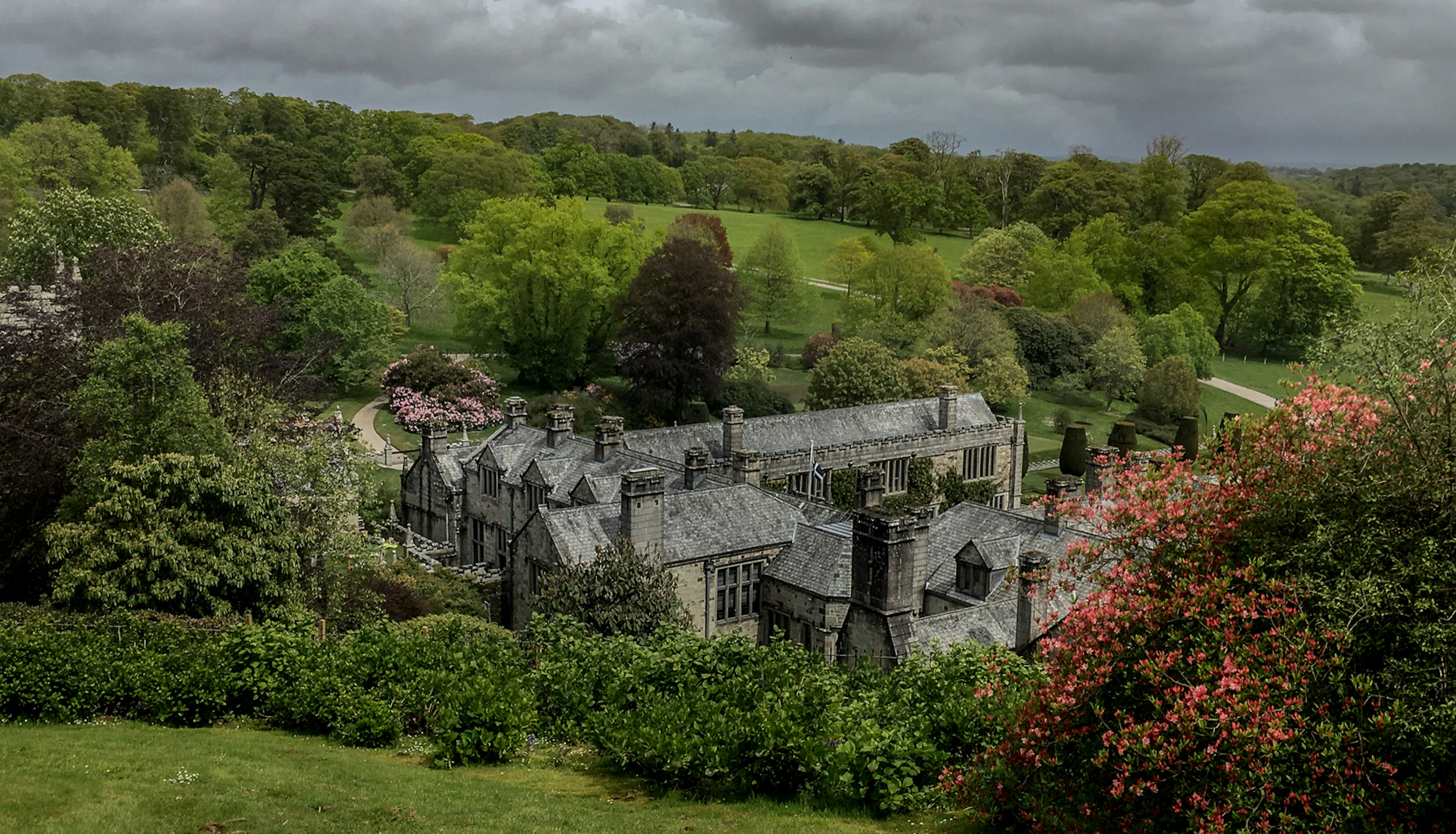 Historic stone manor house surrounded by lush green gardens and trees under a cloudy sky