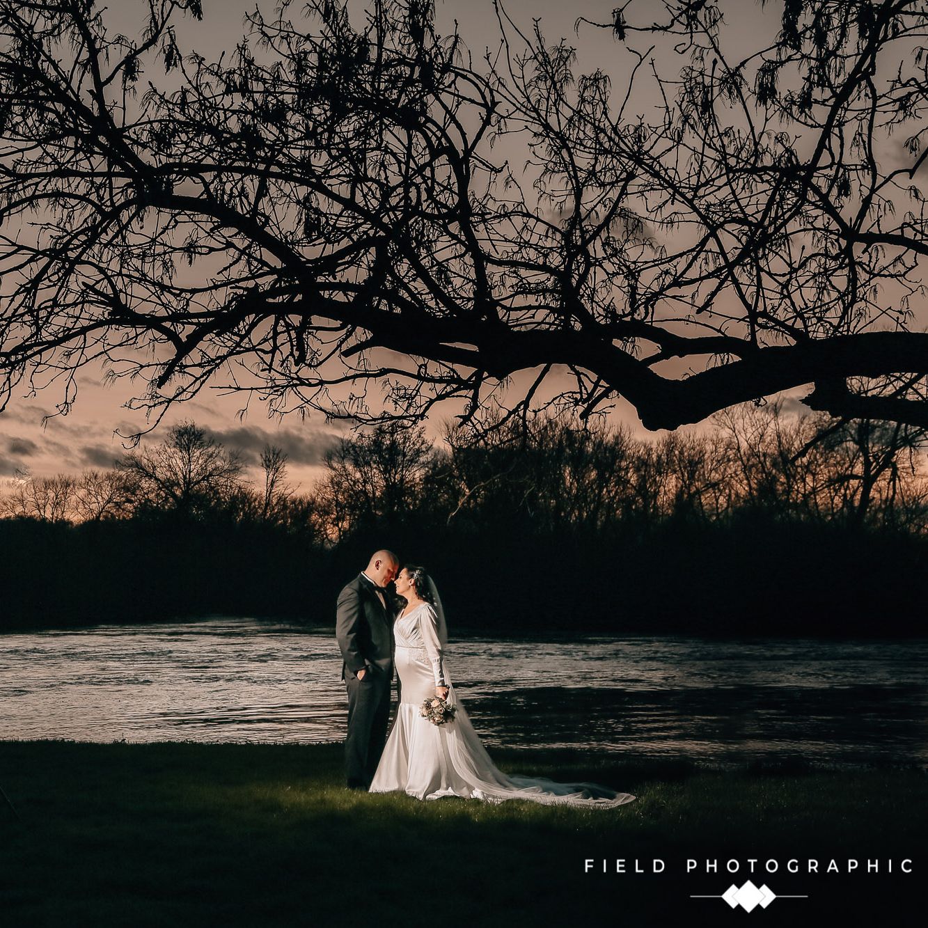 Bride and groom standing together under a large tree by a river at dusk.