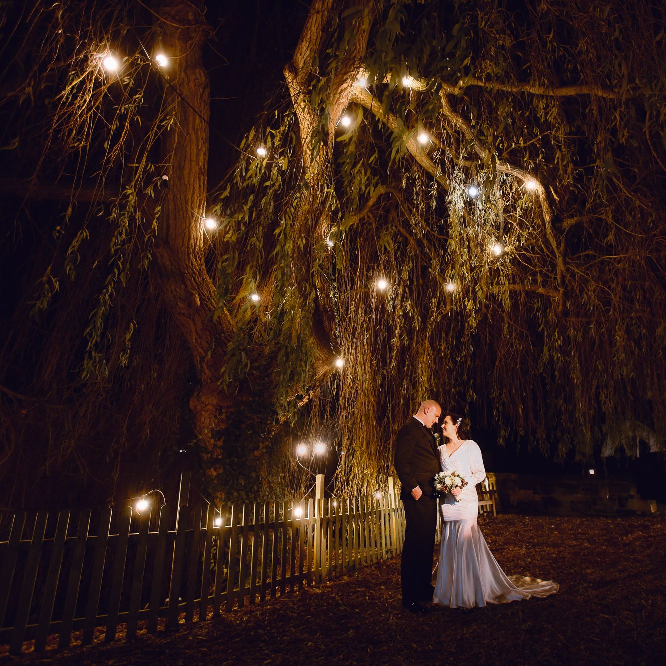 Bride and groom standing under a tree with string lights at night
