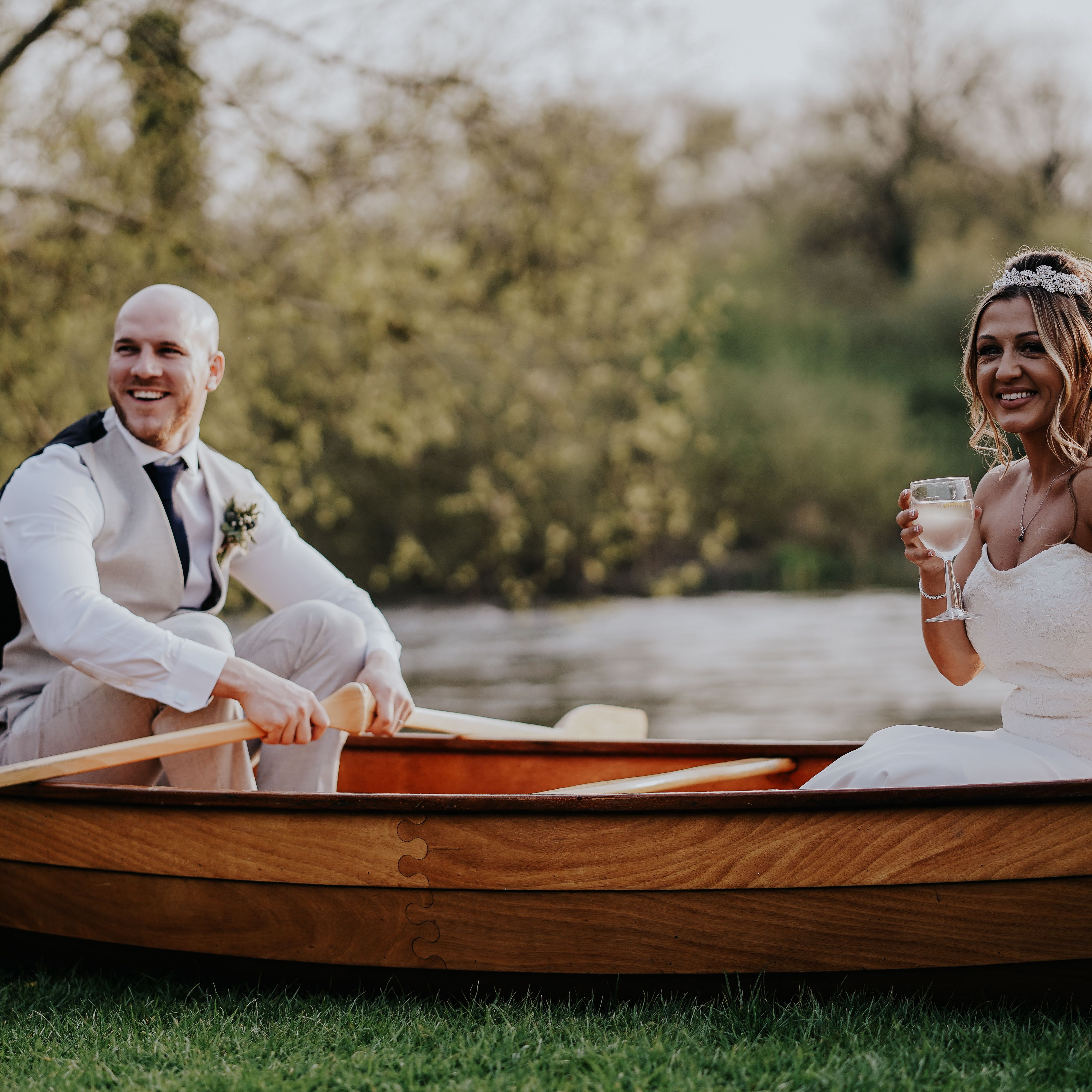 Bride and groom sitting in a wooden boat by a lake, groom rowing and bride holding a glass of wine.