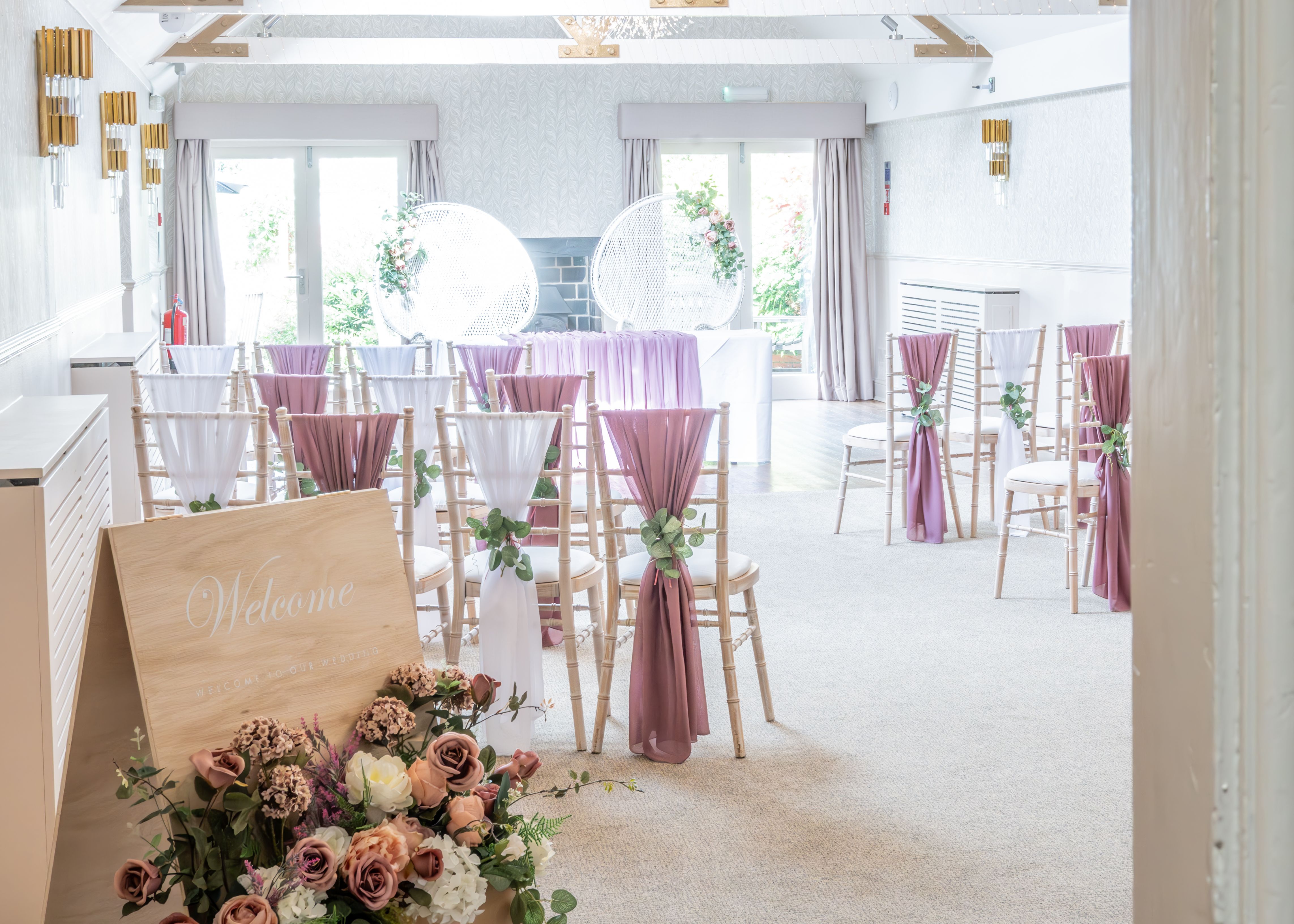 Brightly decorated wedding ceremony room with white ceiling beams, pastel draped chairs, and a welcome sign with flowers