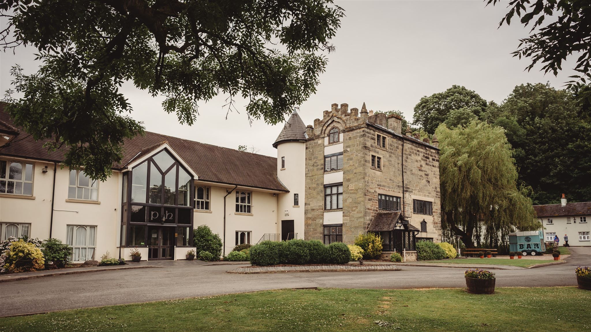 A historic stone manor house with an adjacent modern building, surrounded by greenery and a landscaped lawn.