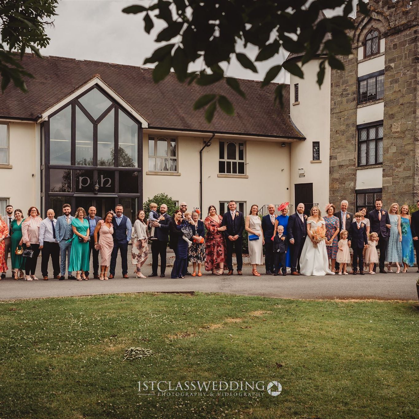 Large group photo of wedding guests standing outside a venue with cream walls and large windows.