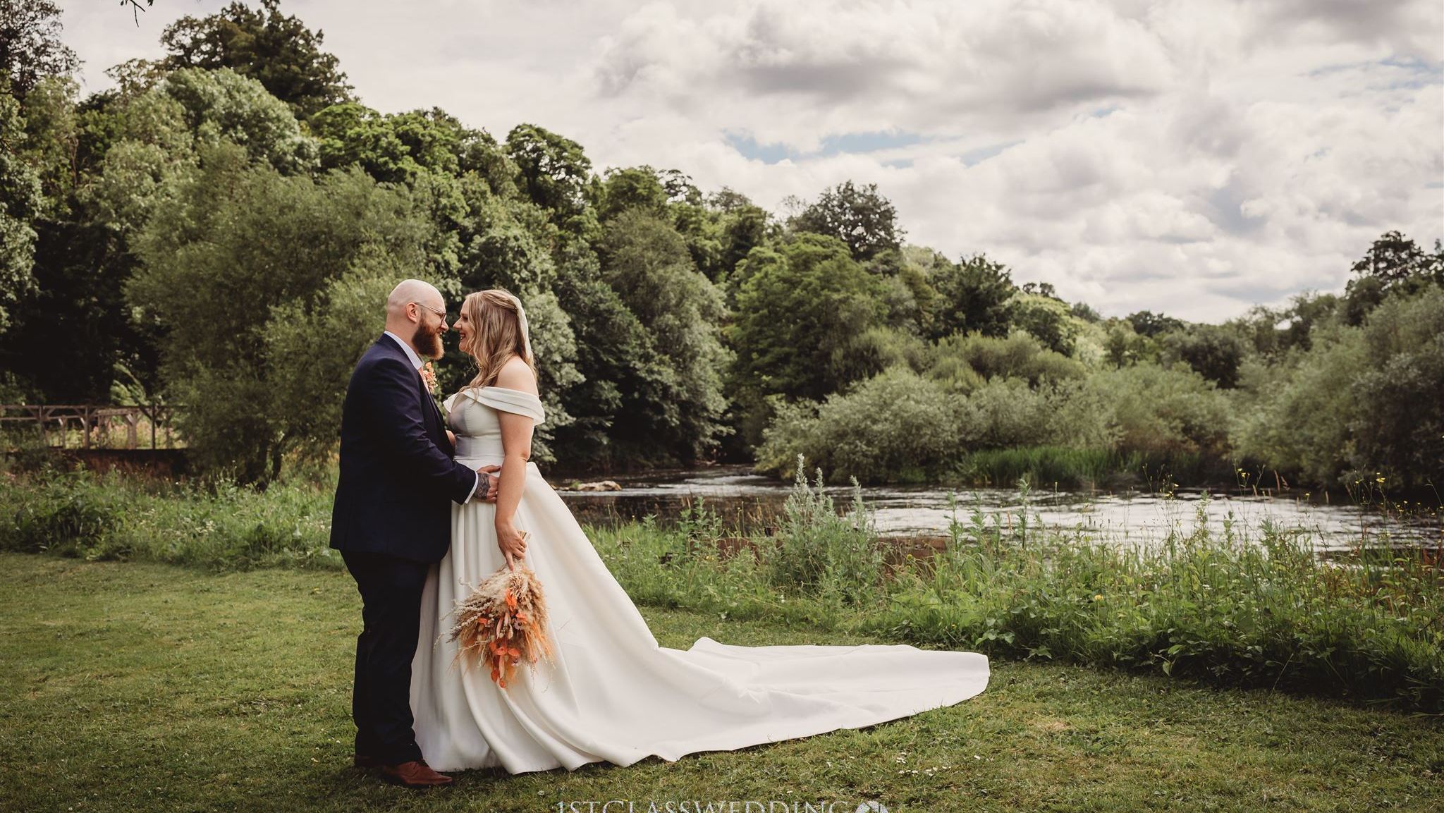 Bride and groom standing together outdoors by a river, surrounded by greenery, on their wedding day.