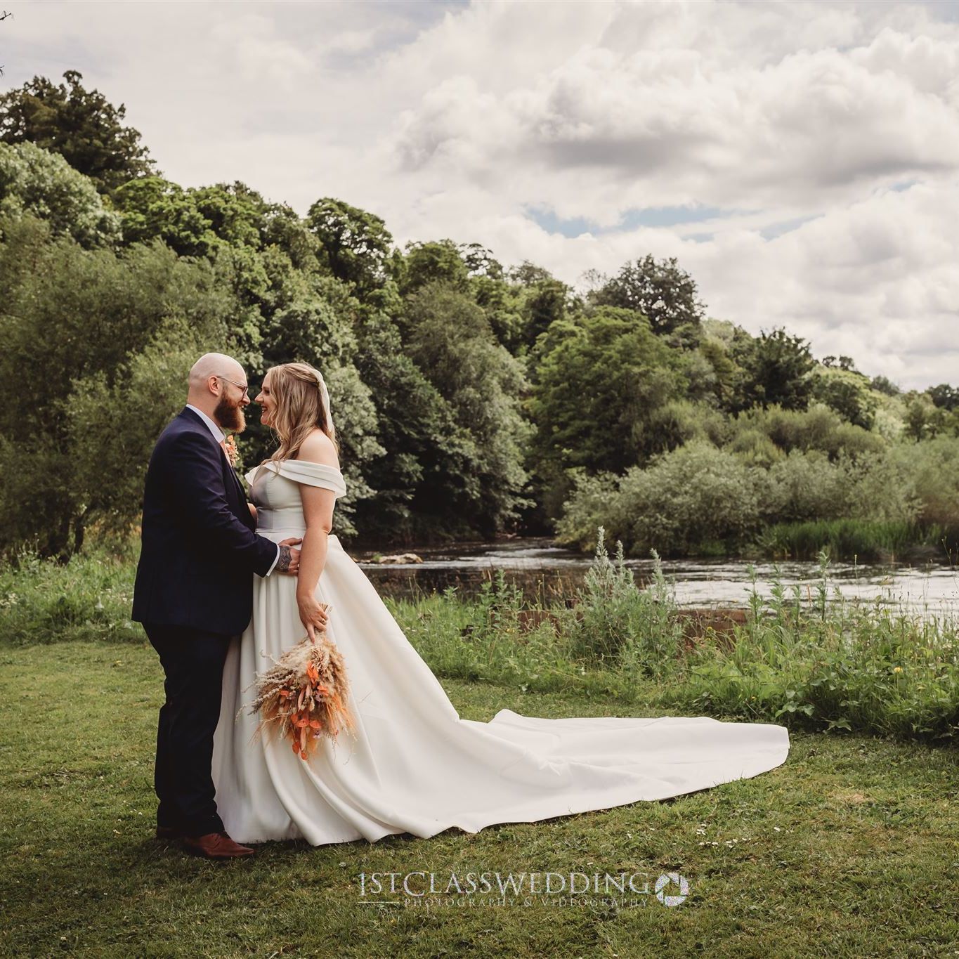 Bride and groom standing together outdoors by a river, surrounded by greenery, on their wedding day.