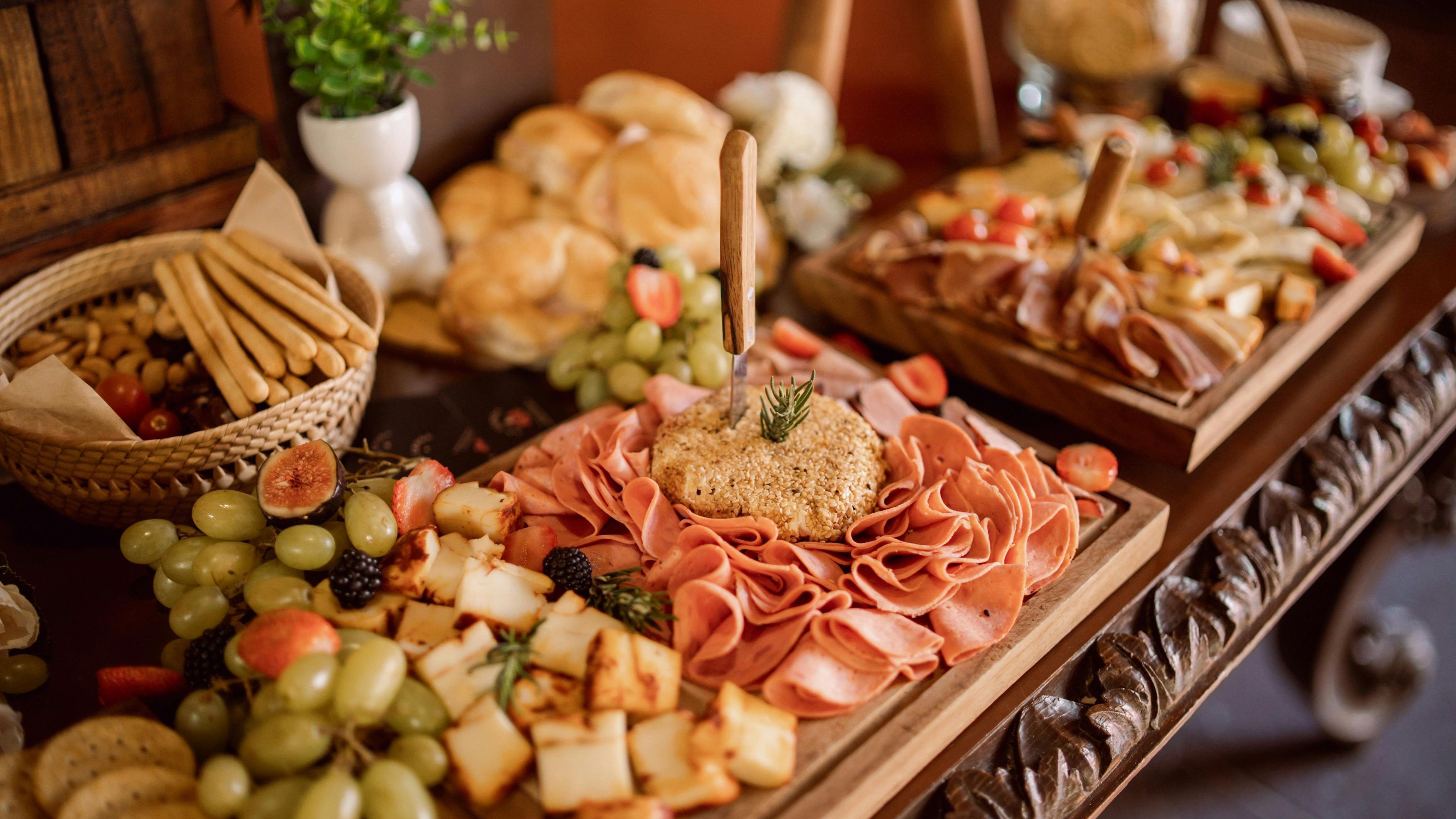 A close-up of a charcuterie board with assorted meats, cheeses, fruits, and a cheese wheel with a knife, surrounded by bread and breadsticks.