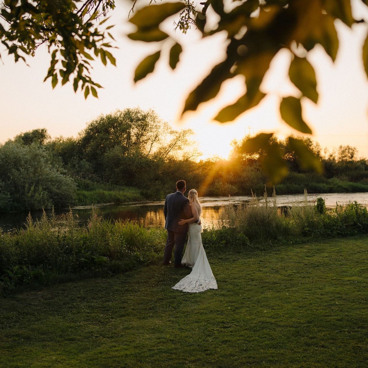 Bride and groom standing by a river at sunset, surrounded by greenery.