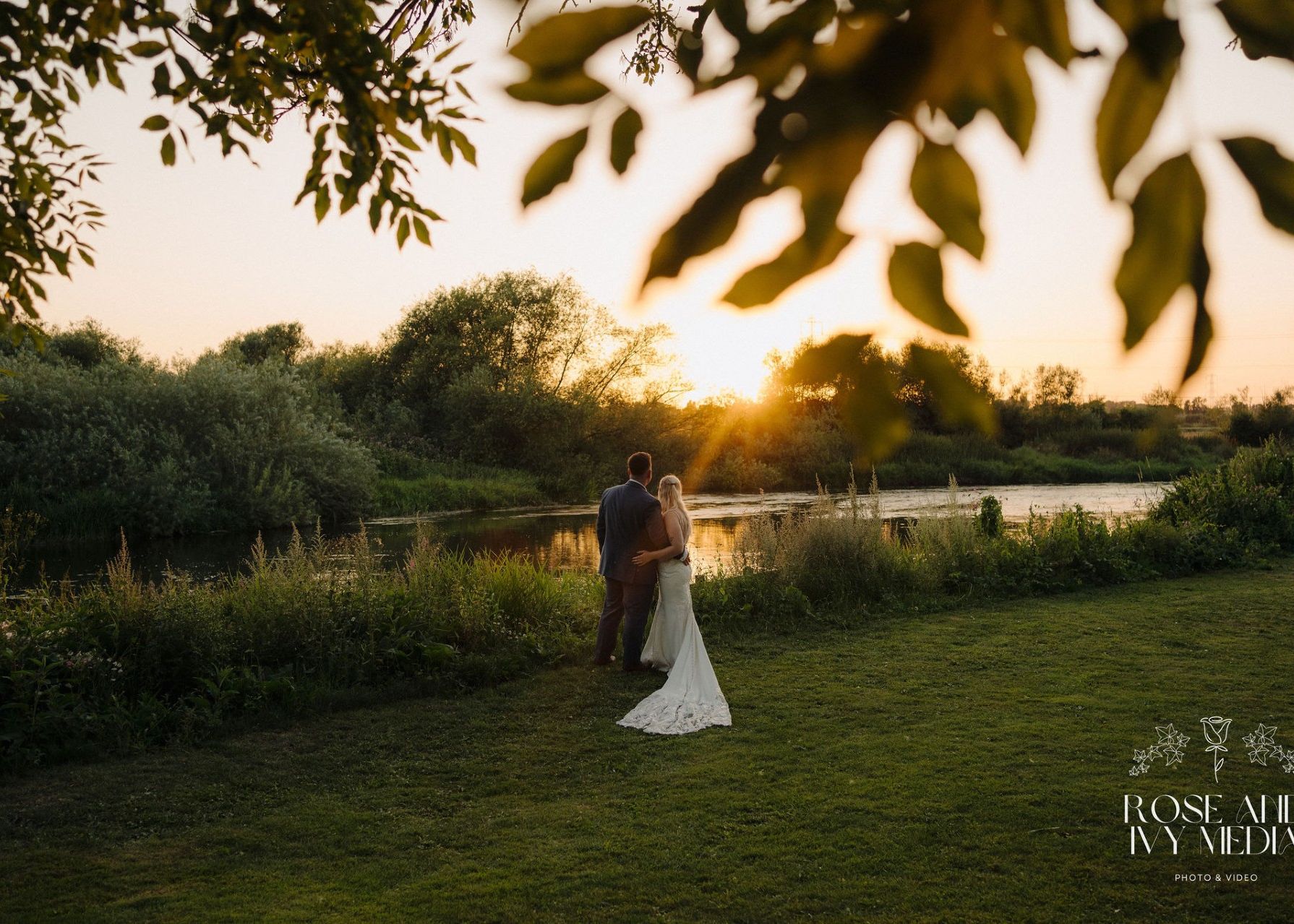 Bride and groom standing by a river at sunset, surrounded by greenery.
