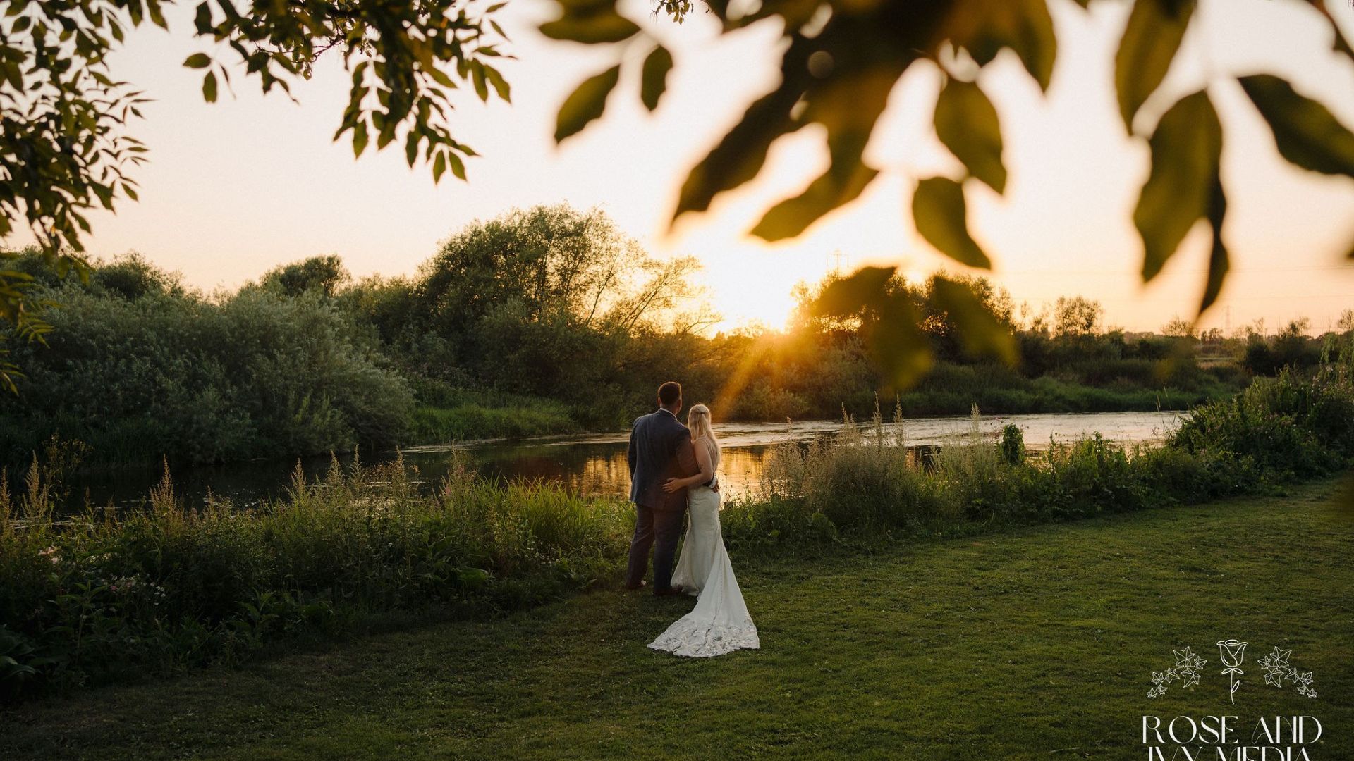 Bride and groom standing by a river at sunset, surrounded by greenery.
