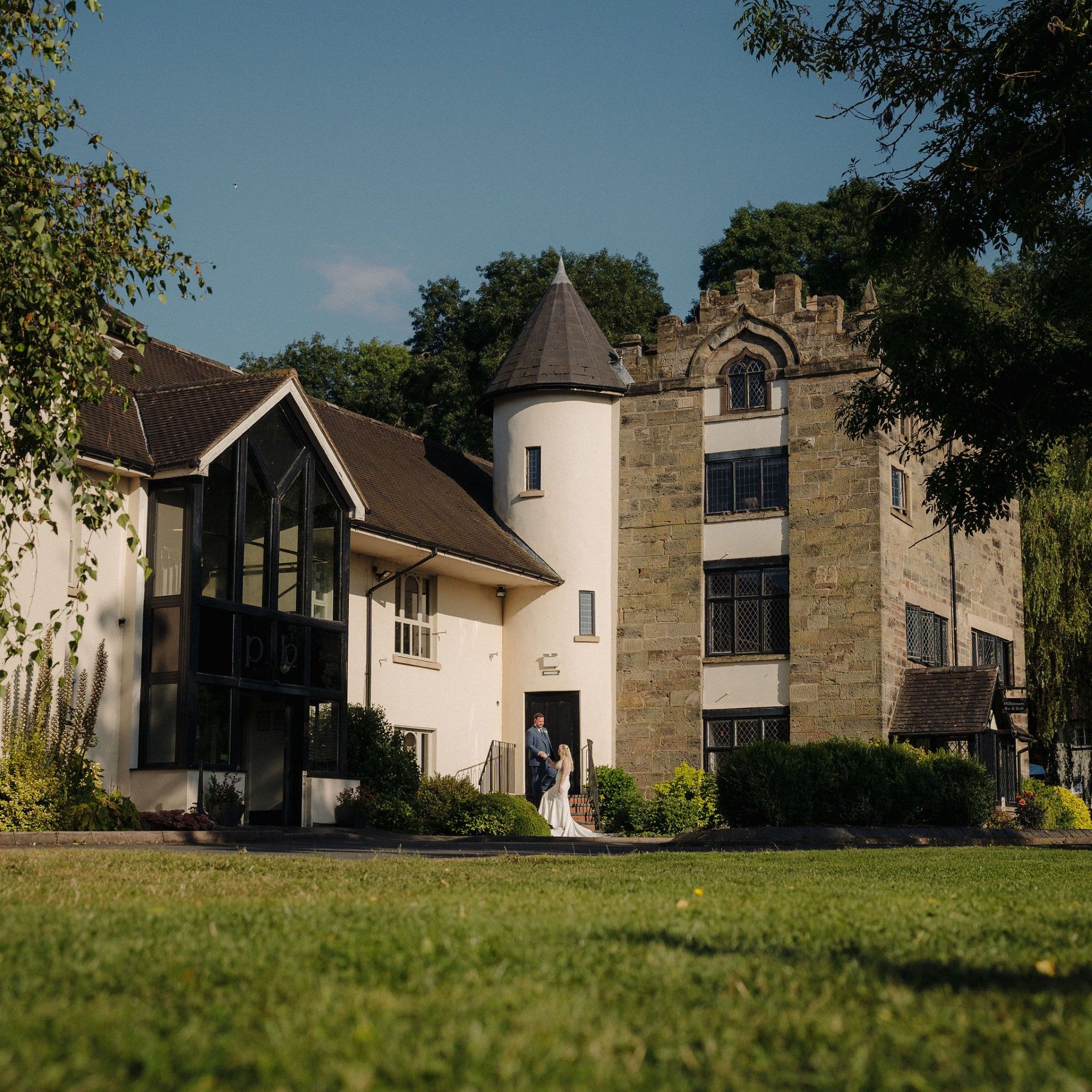 Bride and groom standing outside a historic building with a castle-like tower and modern glass entrance, surrounded by greenery.