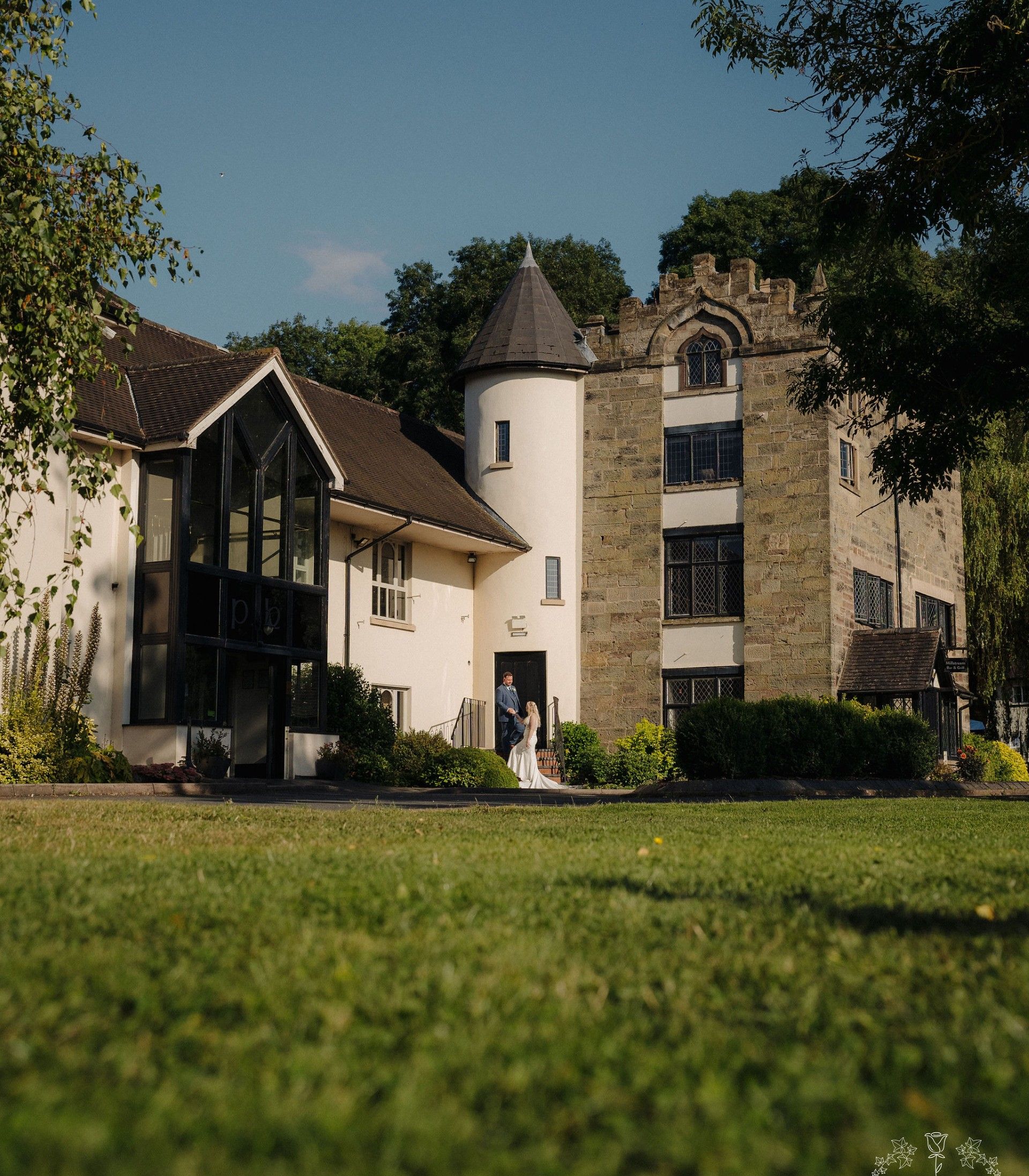 Bride and groom standing outside a historic building with a castle-like tower and modern glass entrance, surrounded by greenery.